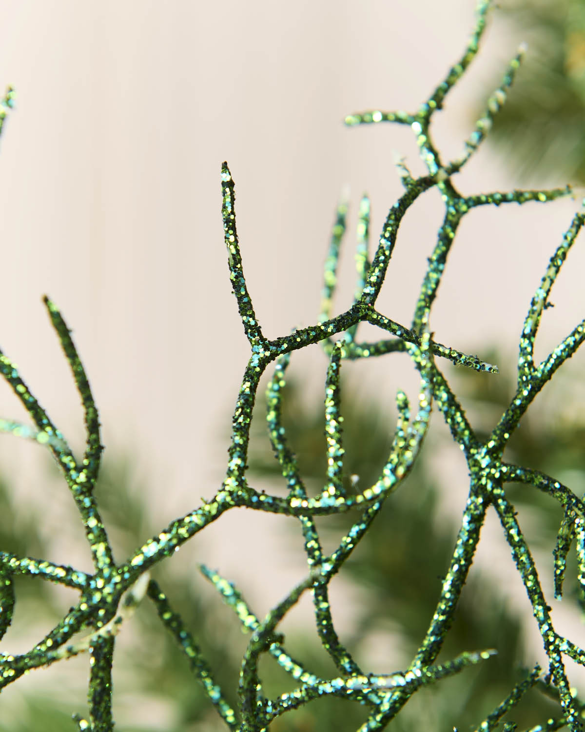 Close-up of a sage green glittery branch-like decorative object with branching, twig-like arms against a soft beige background.