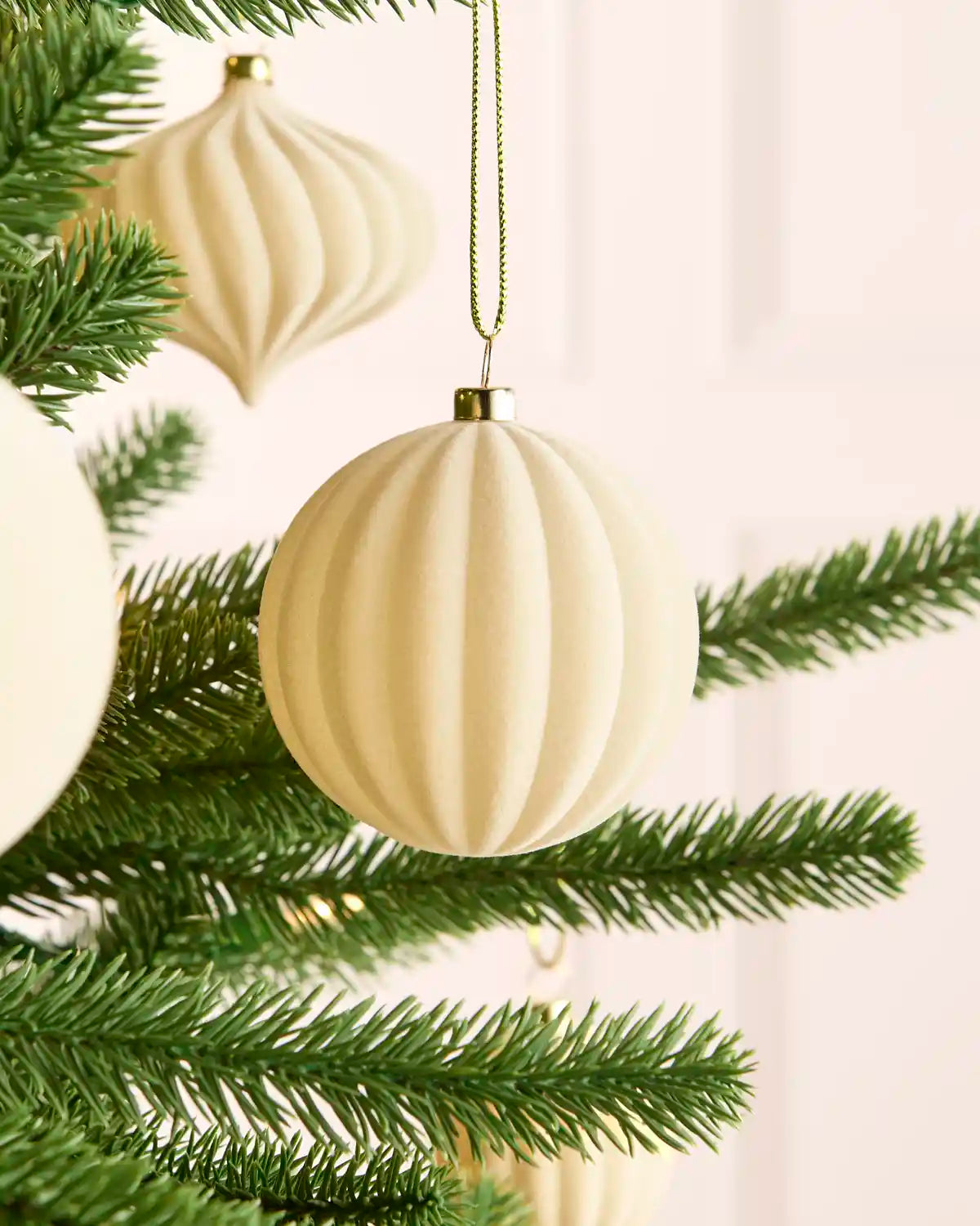 Close-up of a smooth, round latte-coloured velvet bauble hanging from a Christmas tree branch, with another ribbed bauble softly blurred in the background