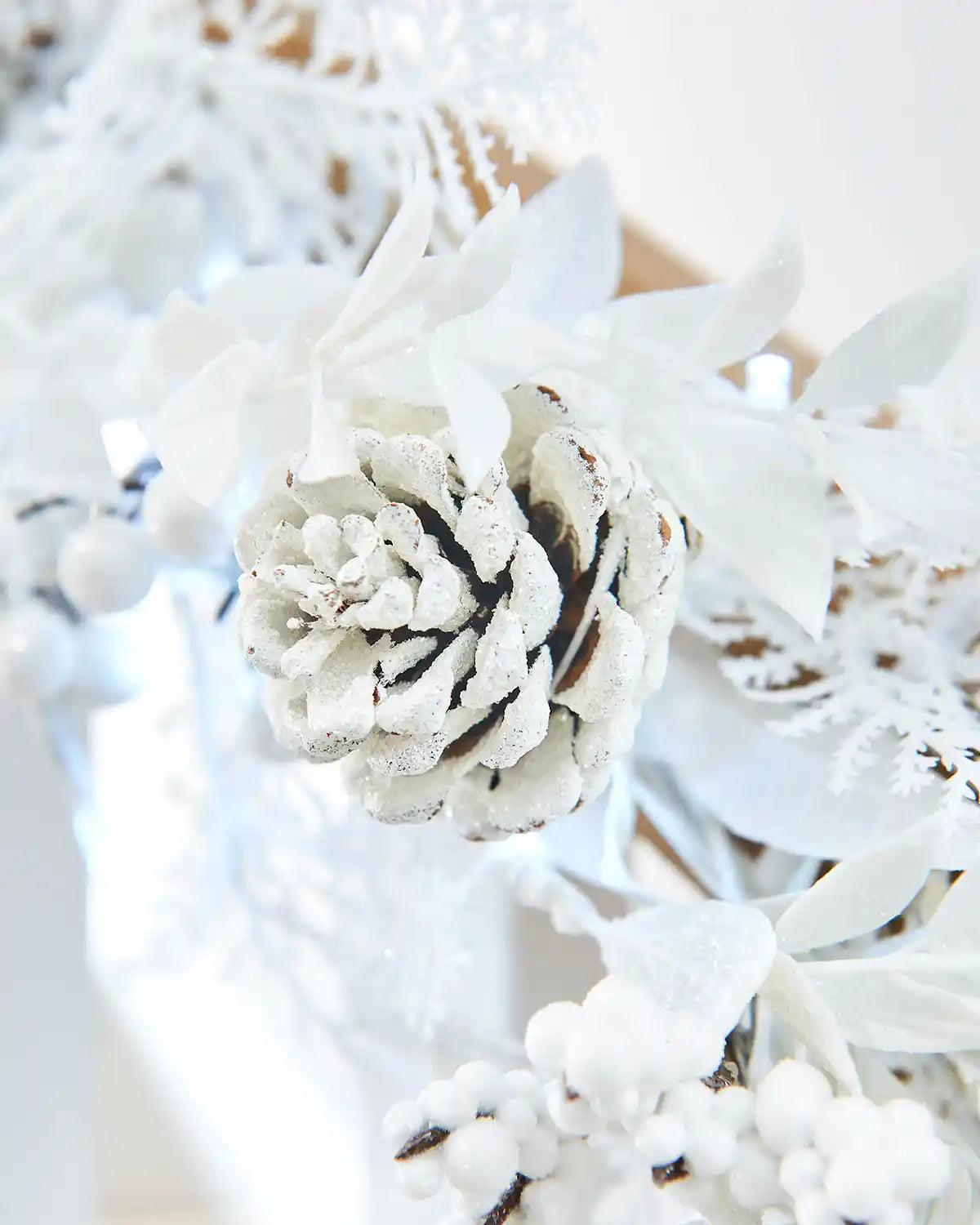Close-up of a white pine cone with snow on it, surrounded by berries and ice white lights 
