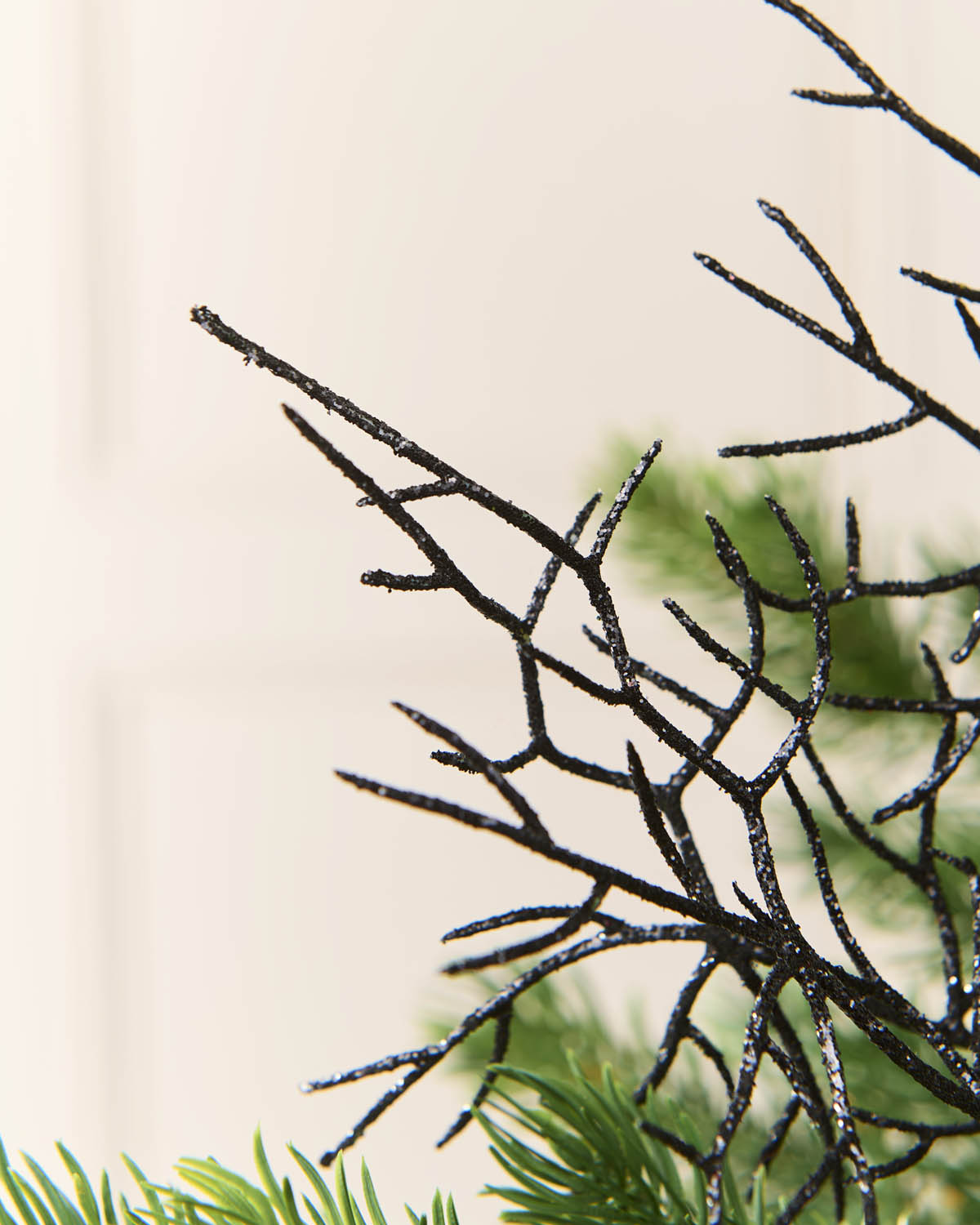 Close-up of black glittery coral style decorative twigs with green pine needles in the foreground against a soft cream background.