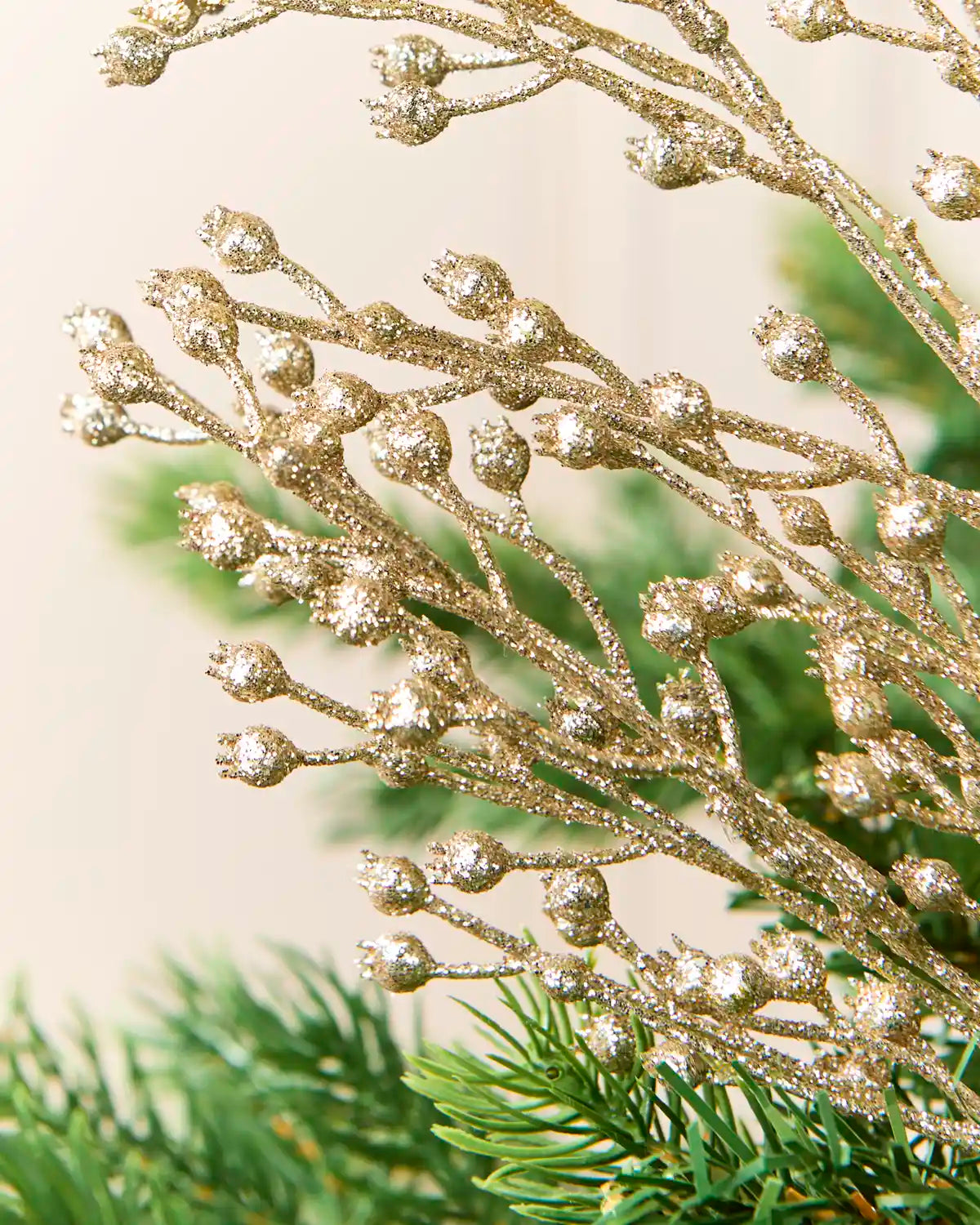 Close-up of glittery gold decorative branches with small round buds, arranged over green pine needles against a soft cream background.