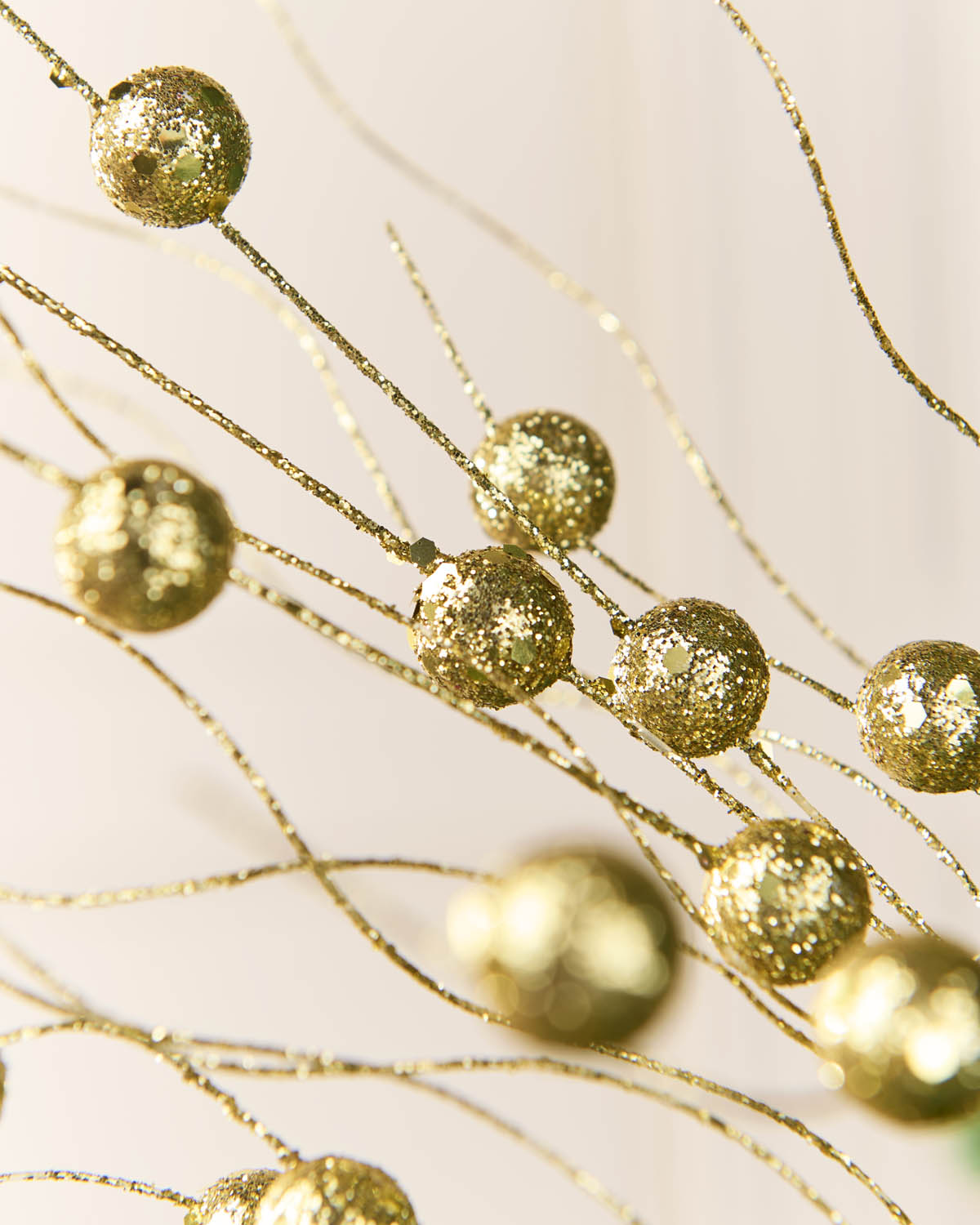 Close-up of gold glitter decorative branches with round metallic beads on thin wire stems against a soft light background.