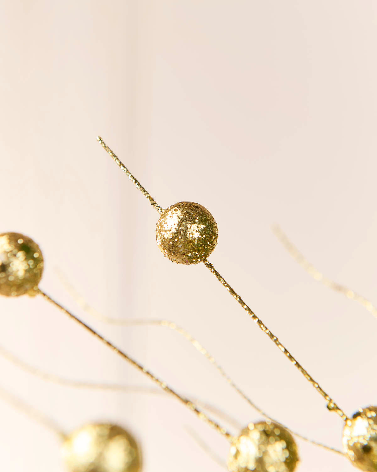 Close-up of gold glitter spherical garland decorations on thin wire stems against a soft warm beige background.