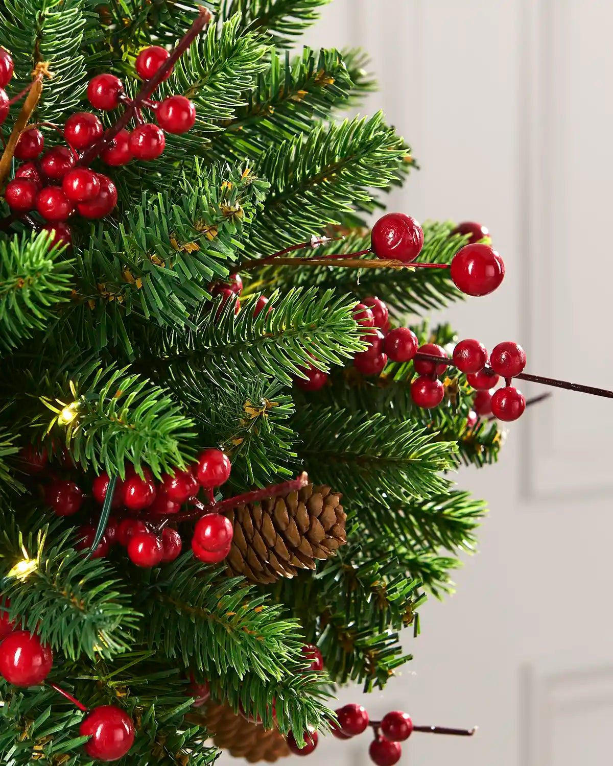 Close-up of green artificial pine branches decorated with glossy red berries, a natural pinecone, and warm white lights