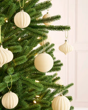 Close-up of latte-coloured velvet baubles hanging from green Christmas tree branches, with warm lights softly glowing in the background