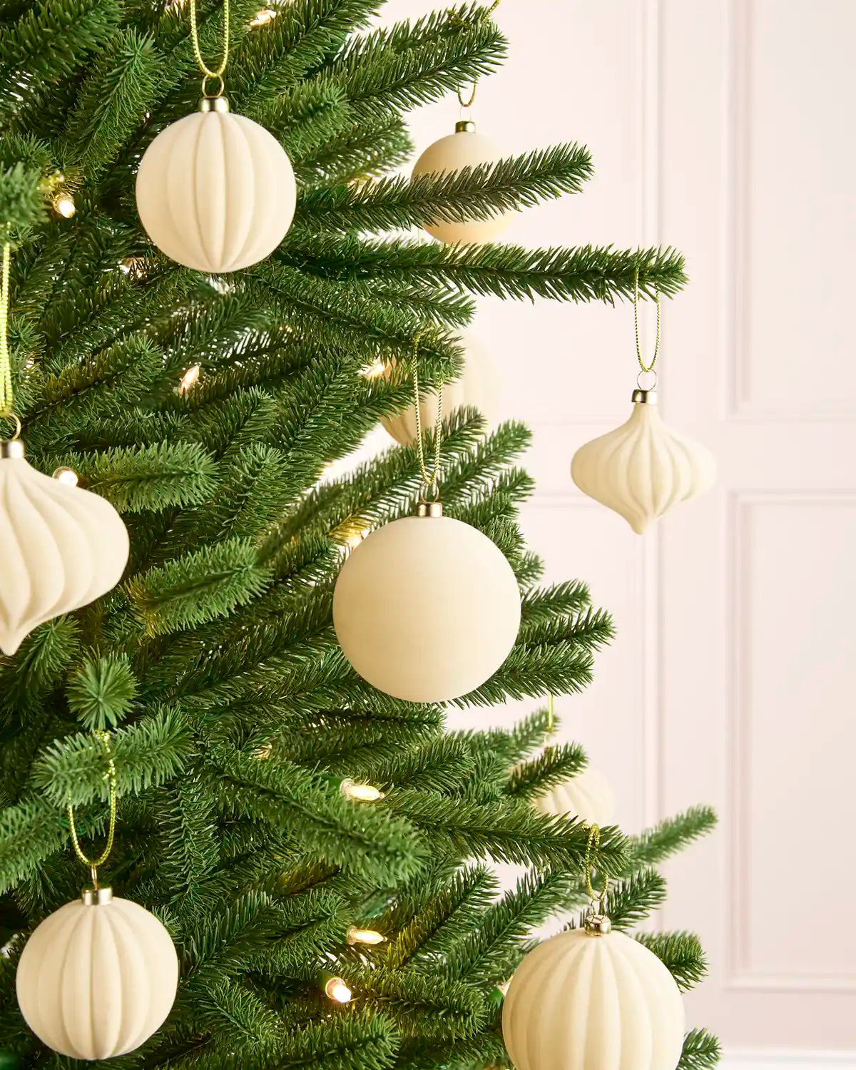 Close-up of latte-coloured velvet baubles hanging from green Christmas tree branches, with warm lights softly glowing in the background