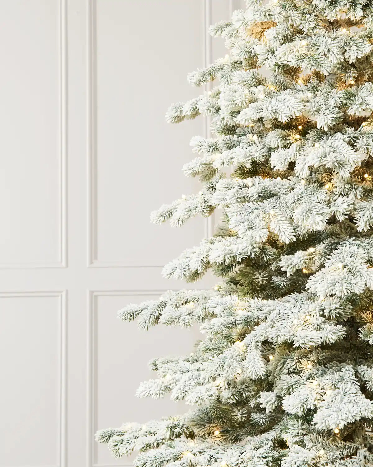 Close-up of snow-covered artificial Christmas tree branches with warm white lights, showcasing the frosted green needles and realistic texture against a white paneled wall.