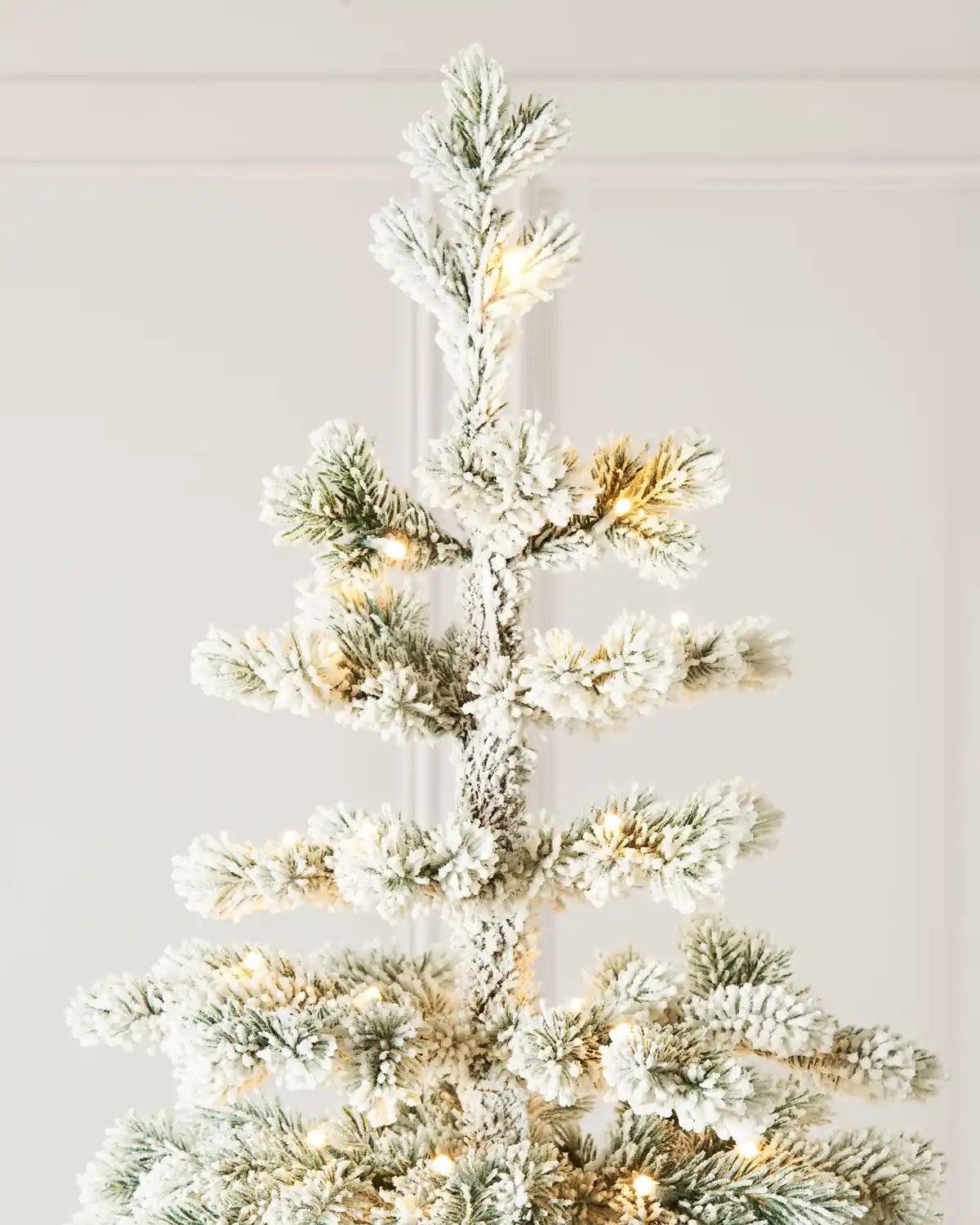 Close-up of the flocked Christmas tree’s top branches, showing the upright central leader, snow-covered needles, and softly glowing warm white lights.