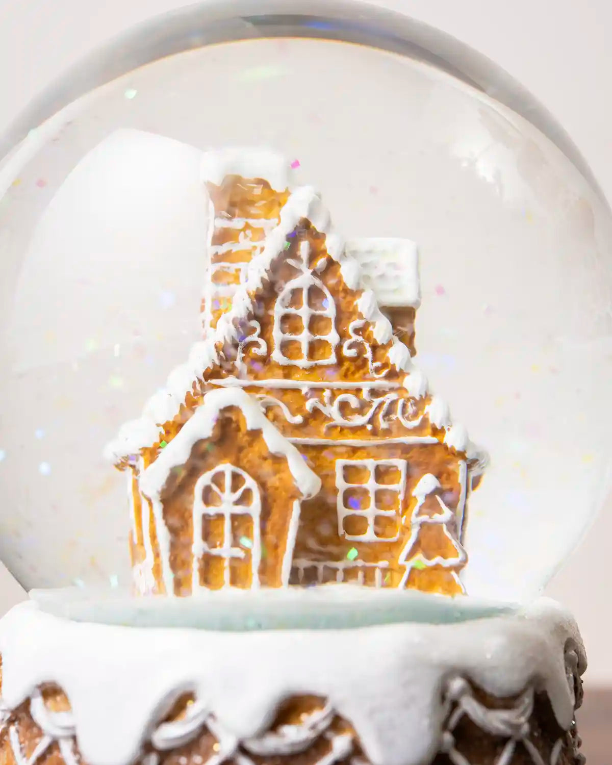 Close-up of the gingerbread house inside a clear snow globe, showing white icing details, windows, a small Christmas tree decoration, and colorful glitter resembling falling snow