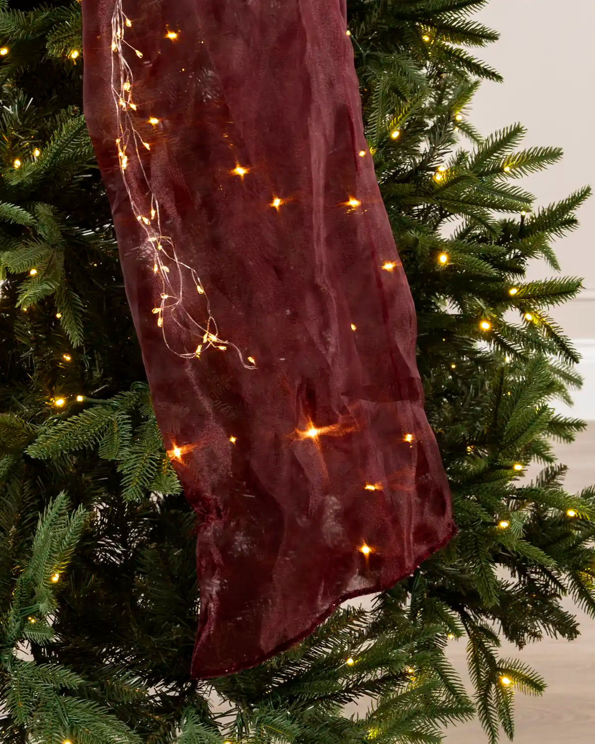 Close-up of the long burgundy bow tail with warm white lights, draped over Christmas tree branches.