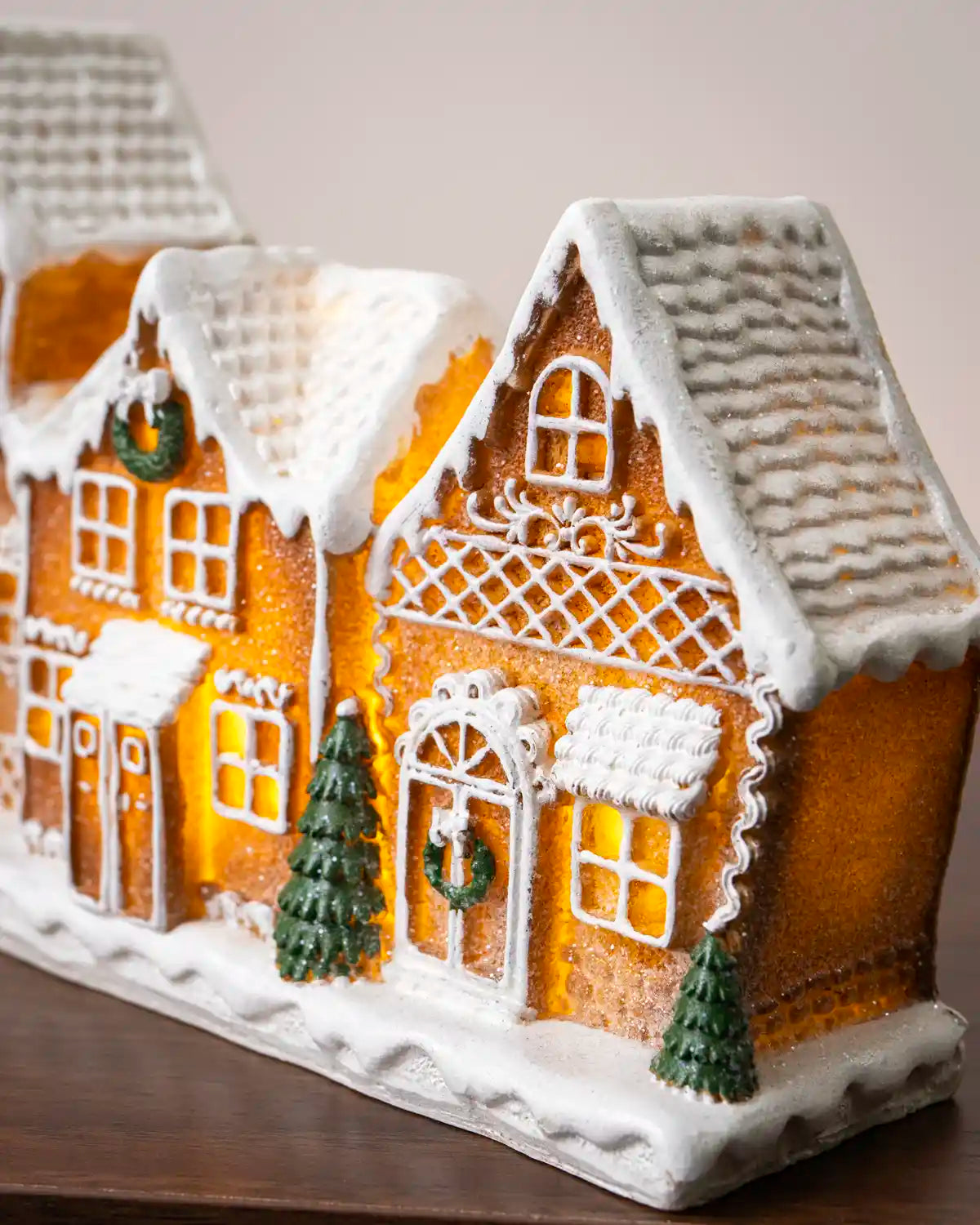 Close-up of the right side of a pre-lit gingerbread village, showing detailed icing decorations, a snow-covered roof, glowing windows, a green wreath, and miniature Christmas trees