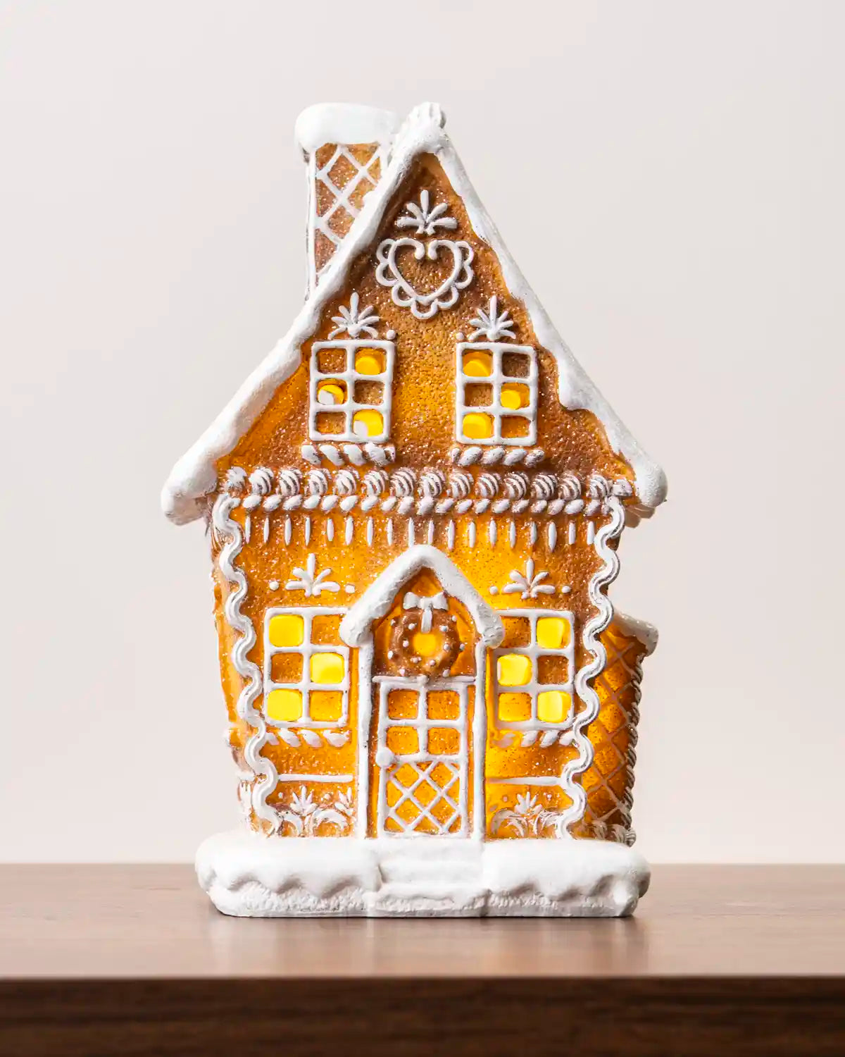 Close-up view of a pre-lit gingerbread house showing its detailed white icing decorations, glowing windows, snowy roof, and front door