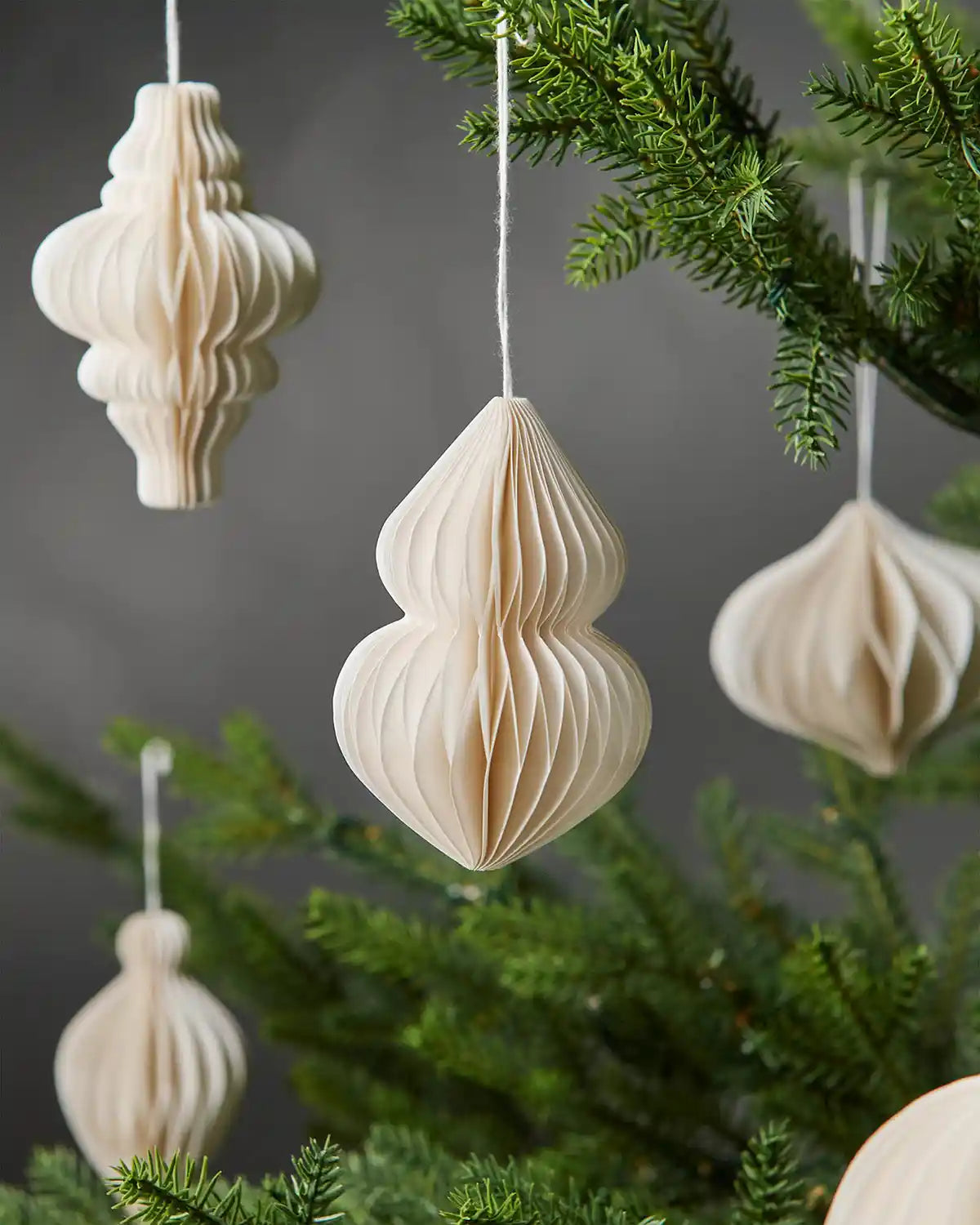 Close-up of 10cm off-white paper Christmas decorations of various shapes hanging on tree in front of grey wall background