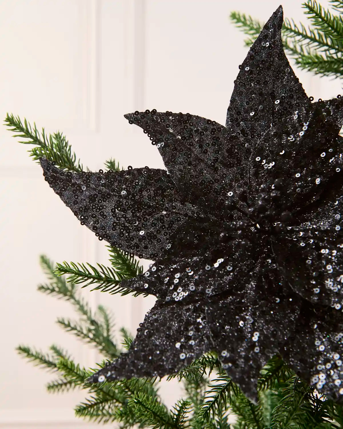 Close up Black Sequin Artificial Poinsettia showing sequin detailing with Christmas tree branches and a cream background
