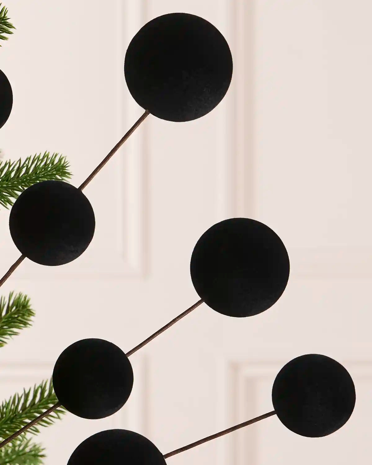 Close up Black velvet bauble Christmas tree pick on thin brown stems, shown hanging against green pine tree branches with a soft white background.
