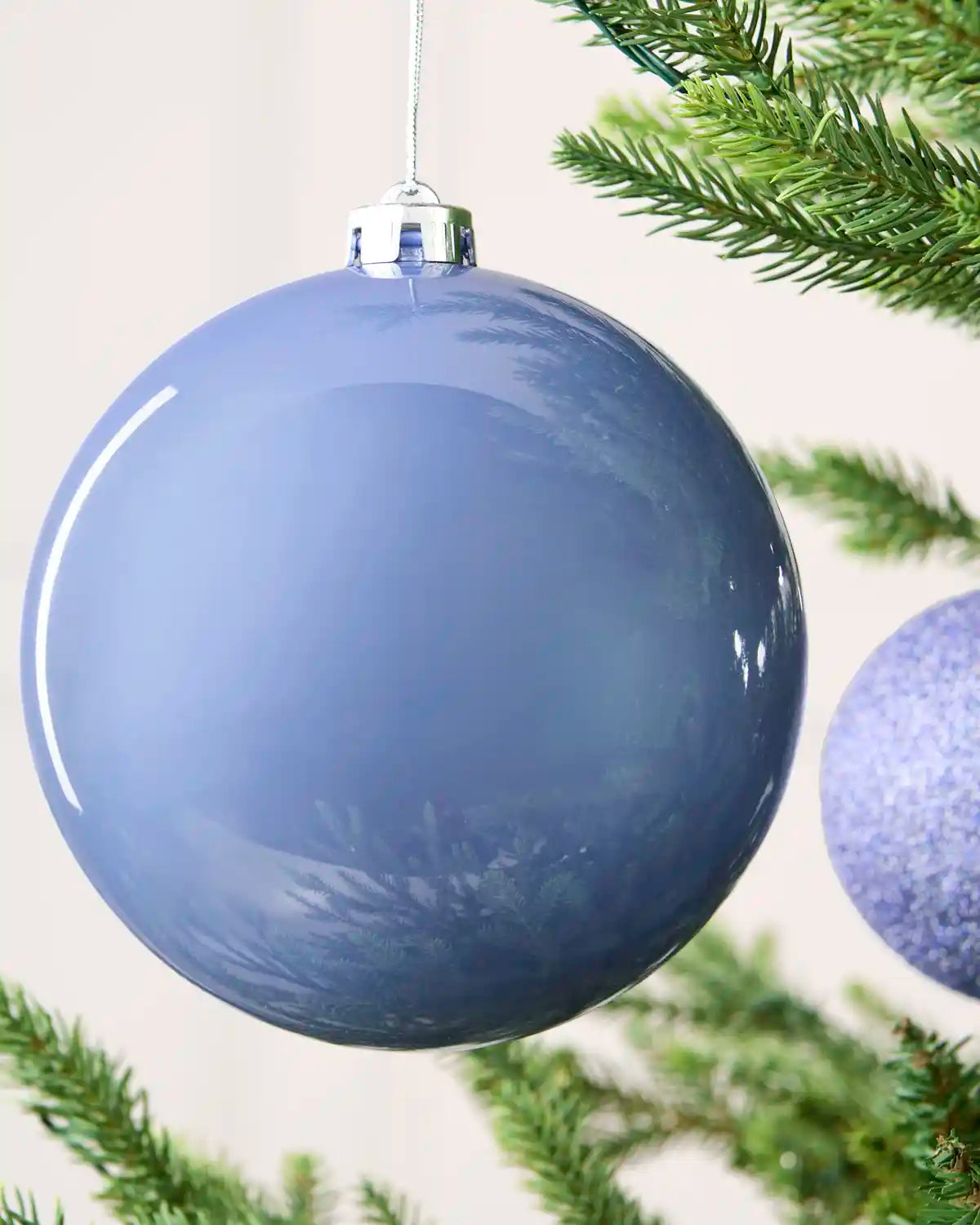 Ice blue shiny oversized bauble with smaller baubles in the background hanging on a Christmas tree against a cream background