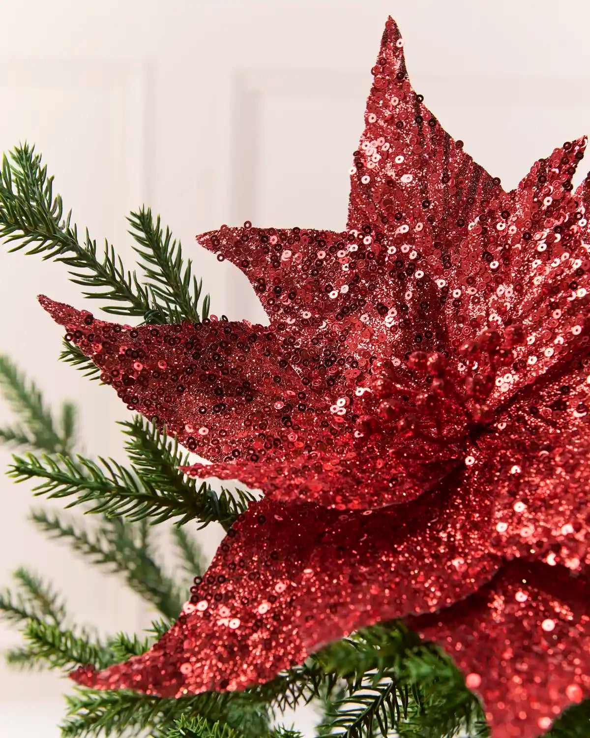 Close up Red Sequin Artificial Poinsettia showing sequin detailing with Christmas tree branches and a cream background