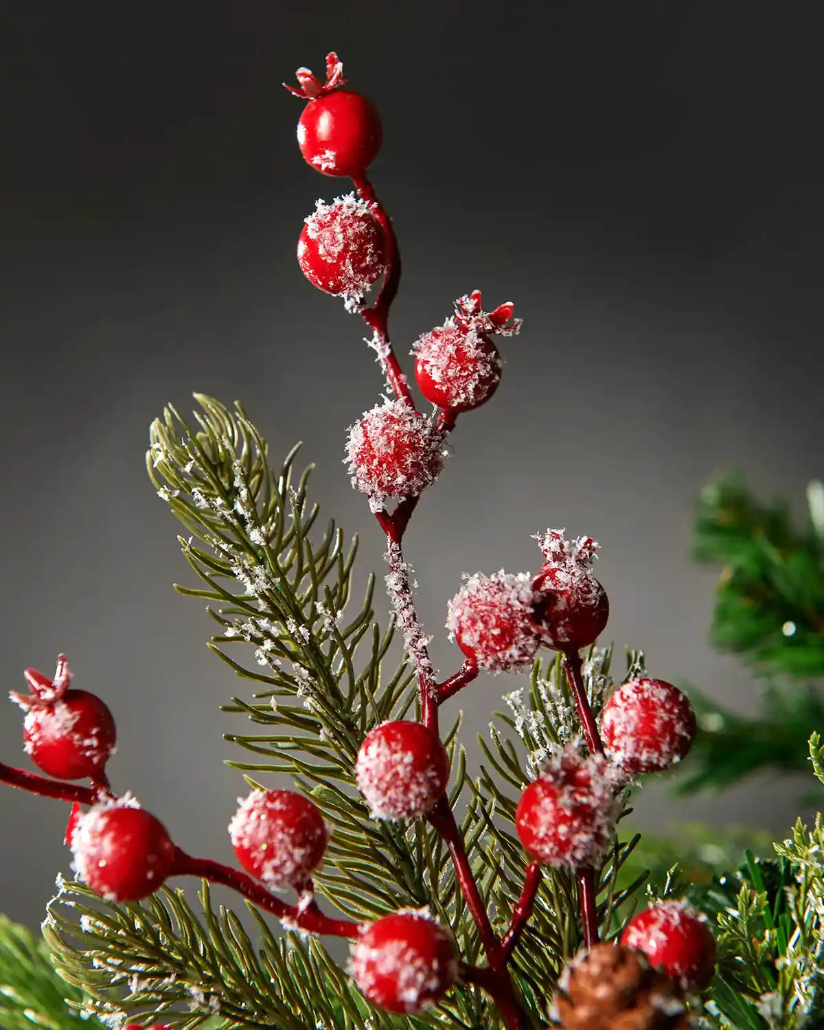 Close-up of the frosted red berries and pinecone pick attached to Christmas tree in front of grey wall background highlighting berry detail