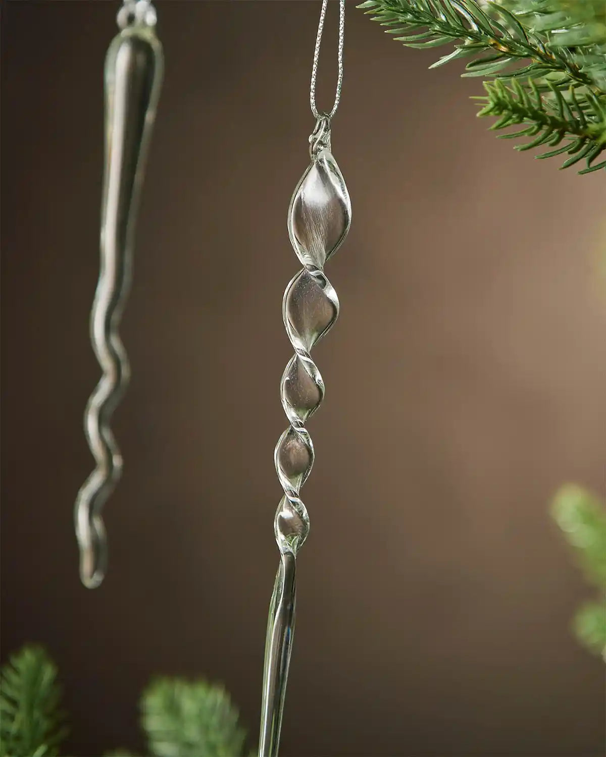 Close up of 2 Decorative glass icicle ornaments hanging from a Christmas tree branch against a brown background