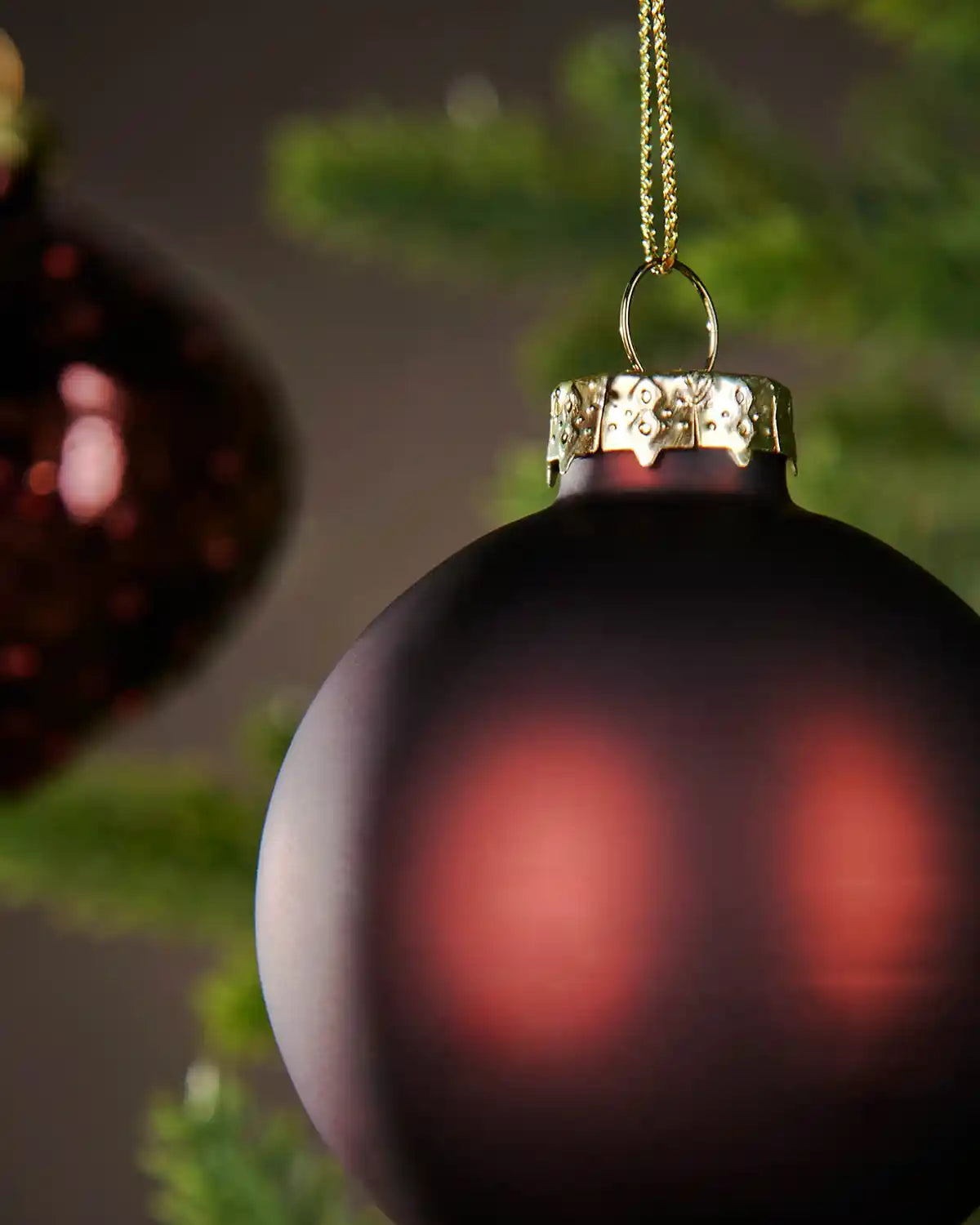 Close up of 2 burgundy baubles hanging on a Christmas tree with a blurry green and brown background