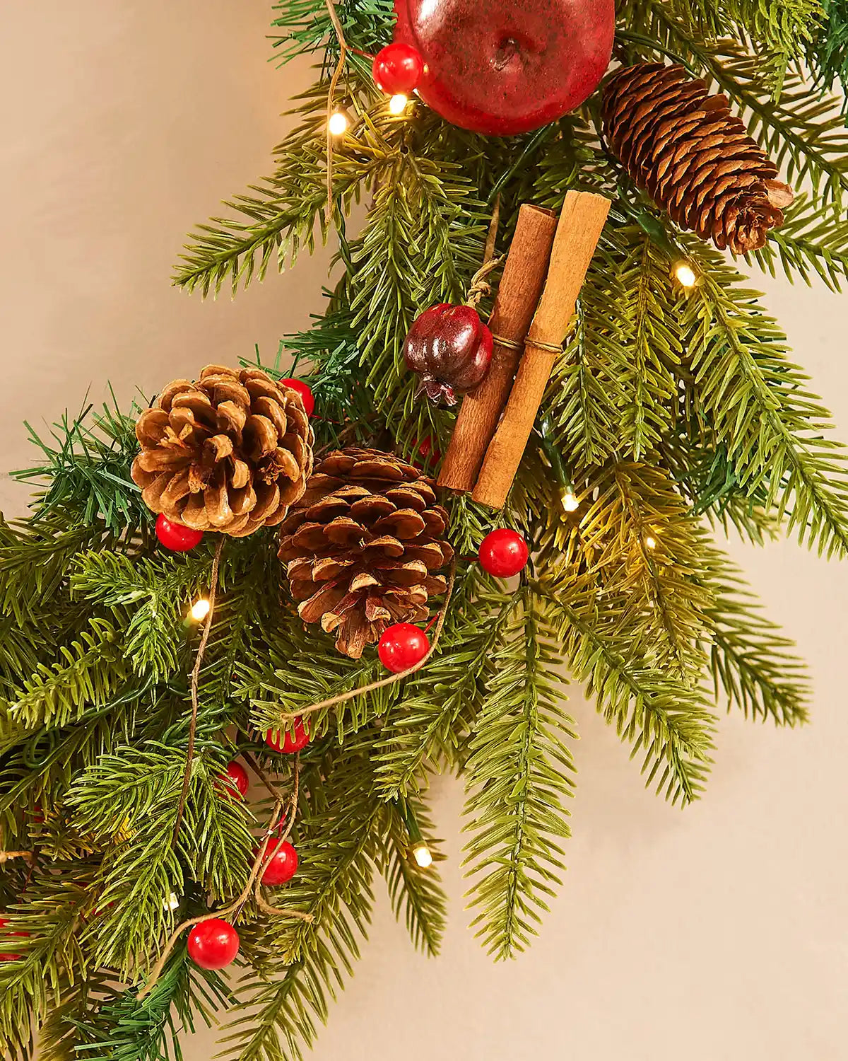 Close up of Apples, cinnamon sticks, pinecones and string lights on a green wreath against a cream wall