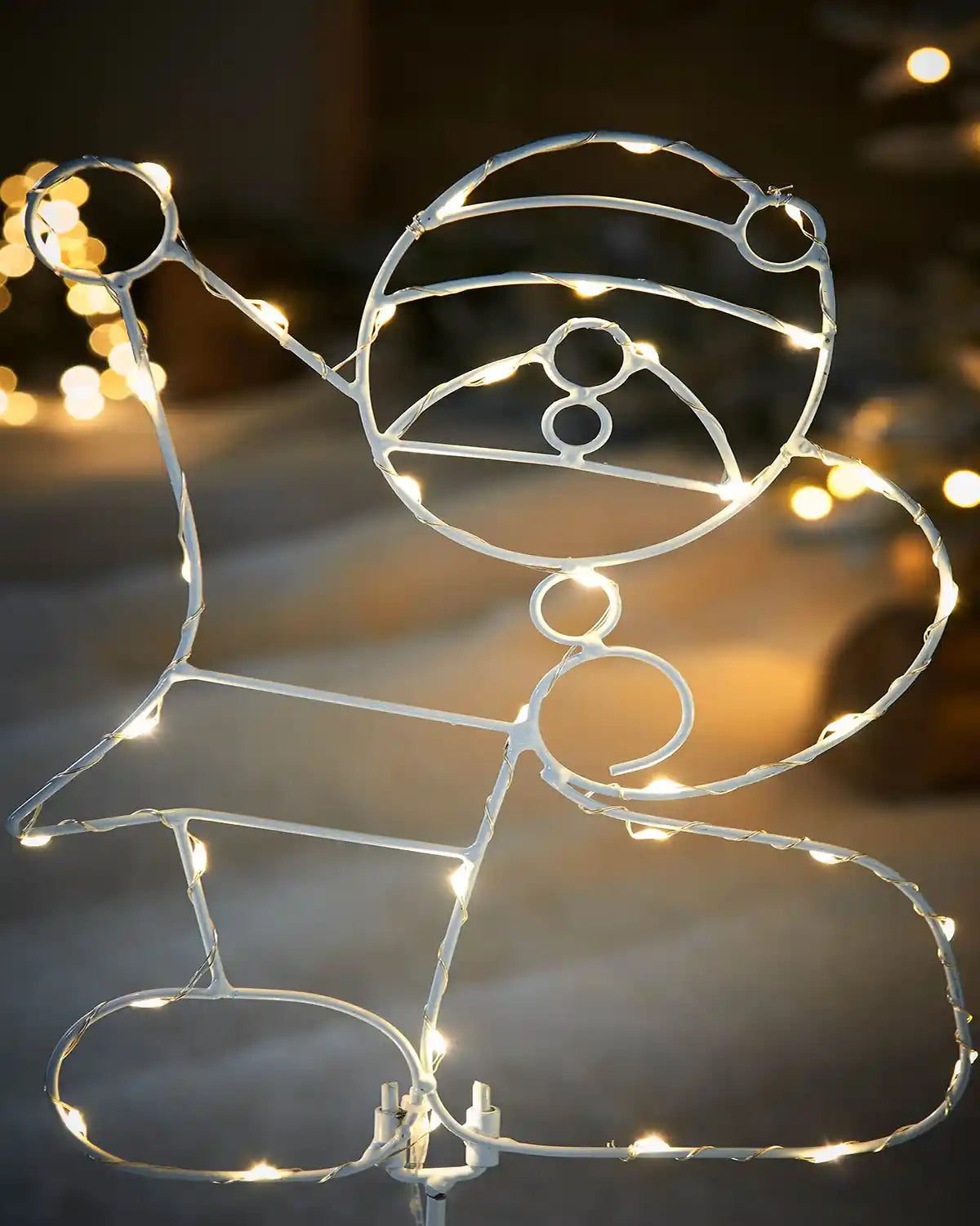 Close up of Decorative wire figure of Santa Claus with a hat and sack, illuminated by string lights against a blurred festive background.