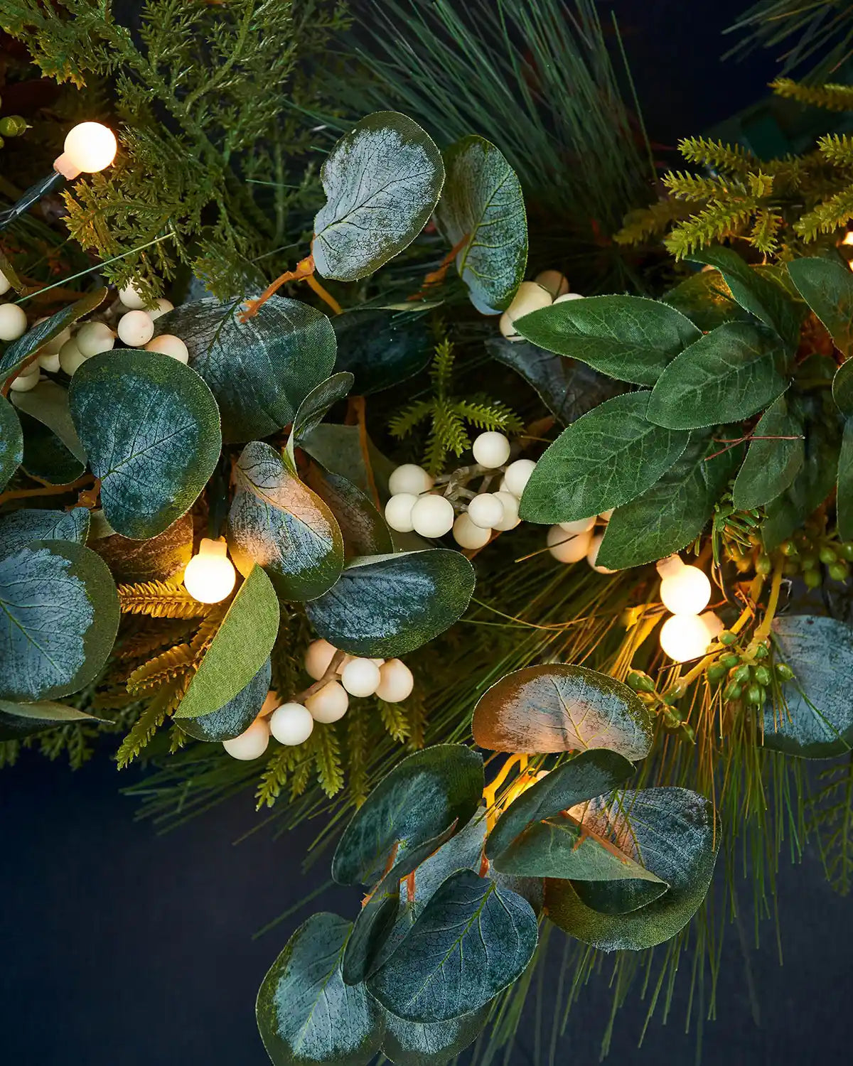 Close up of Decorative wreath with green leaves, white berries, and White berry lights on a dark background