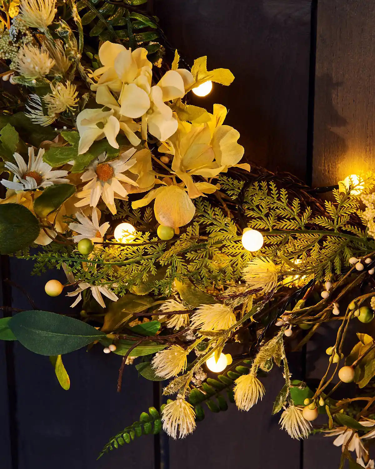 Close up of Decorative wreath with yellow flowers and greenery, illuminated by small lights against a dark background.