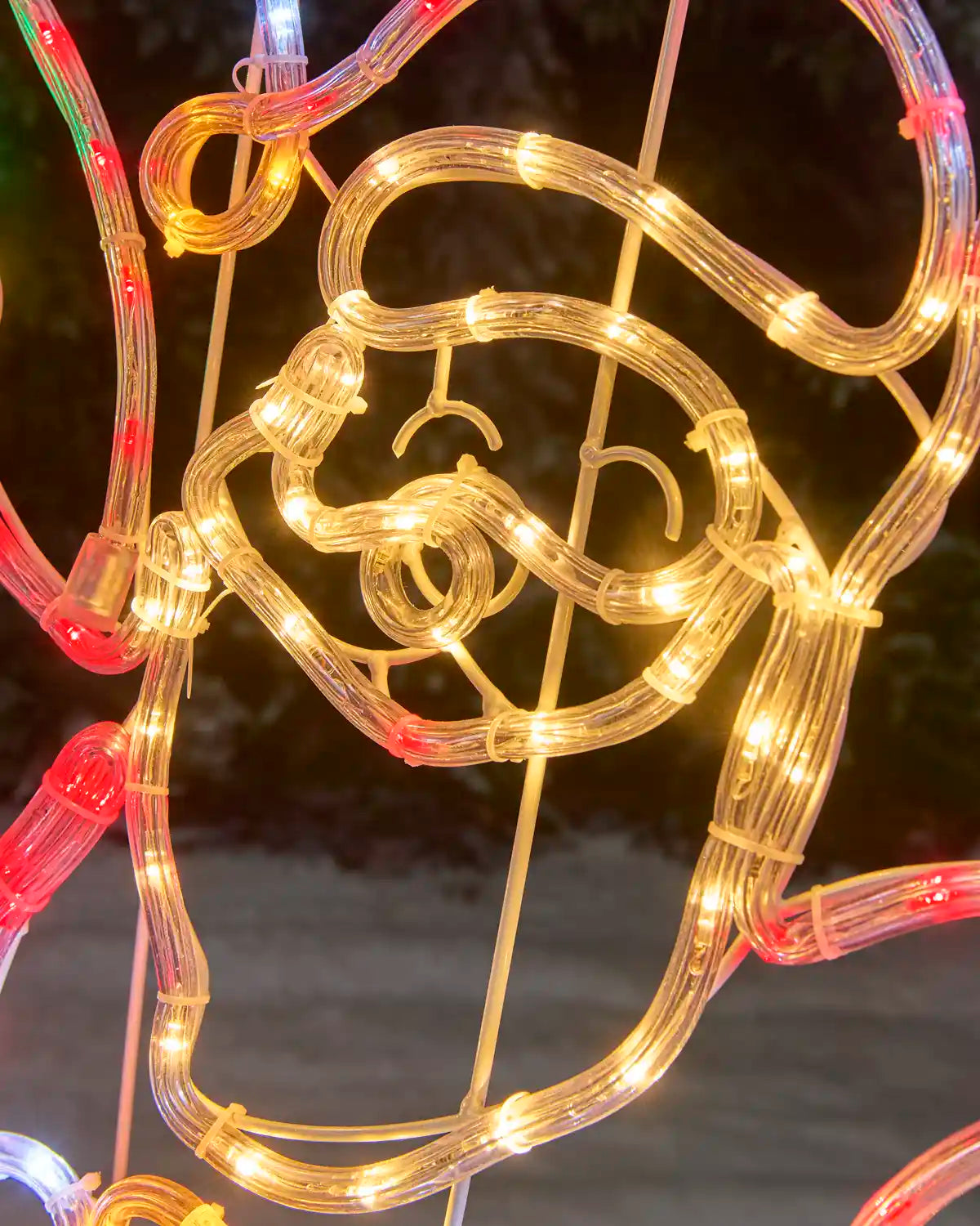 Close up of Santa Clause face in a warm white rope light against a snowy background and dark foliage 