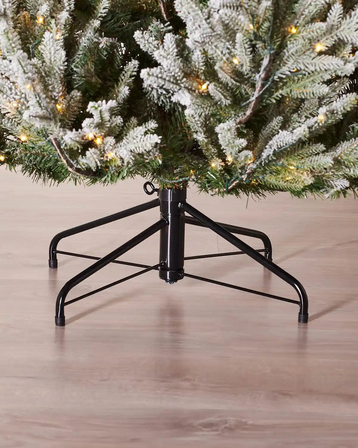 Close up of a Black metal tree stand holding an a snow flocked 8ft  Douglas Fir Christmas tree on a wooden floor.
