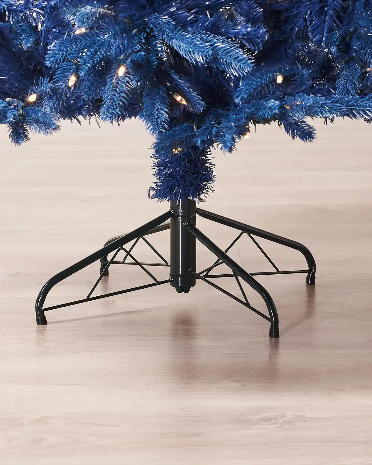 Close up of a Sapphire Blue Christmas tree with a black metal stand on a light wooden floor.