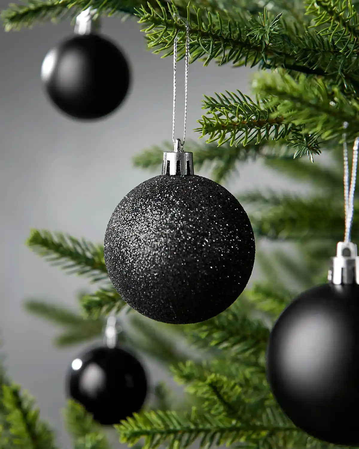 Close up of a glitter black small bauble on a Christmas tree with blurry baubles in the background against a grey wall