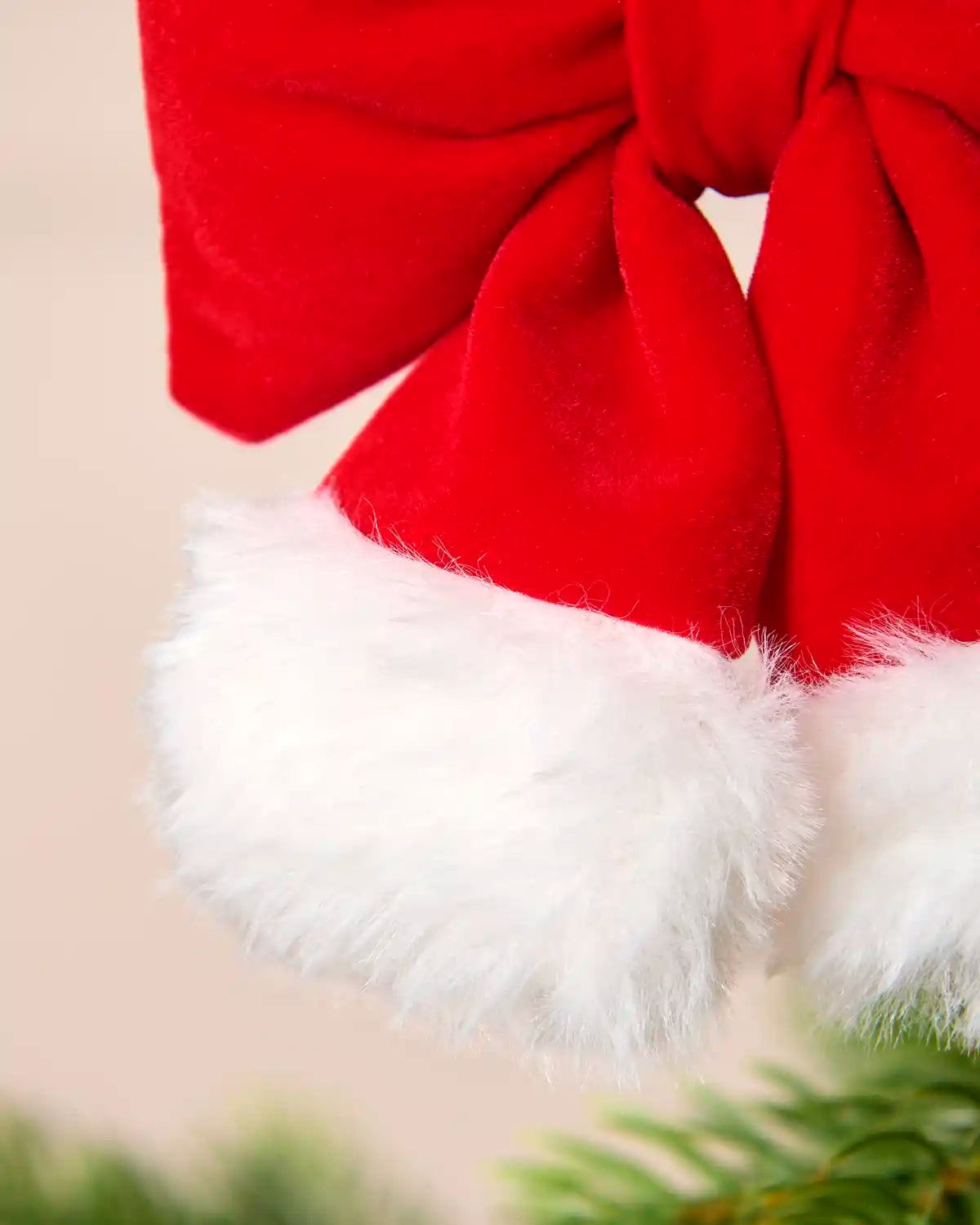 Close up of a red velvet Santa hat ornament shaped like a bow with white faux fur trim, hanging on a green Christmas tree.