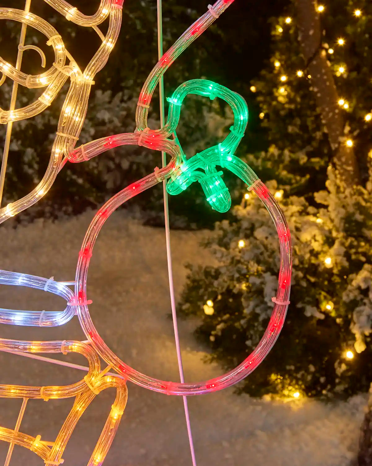 Close up of a rope light Santa holding a present bag against snowy grass and trees with lights on 