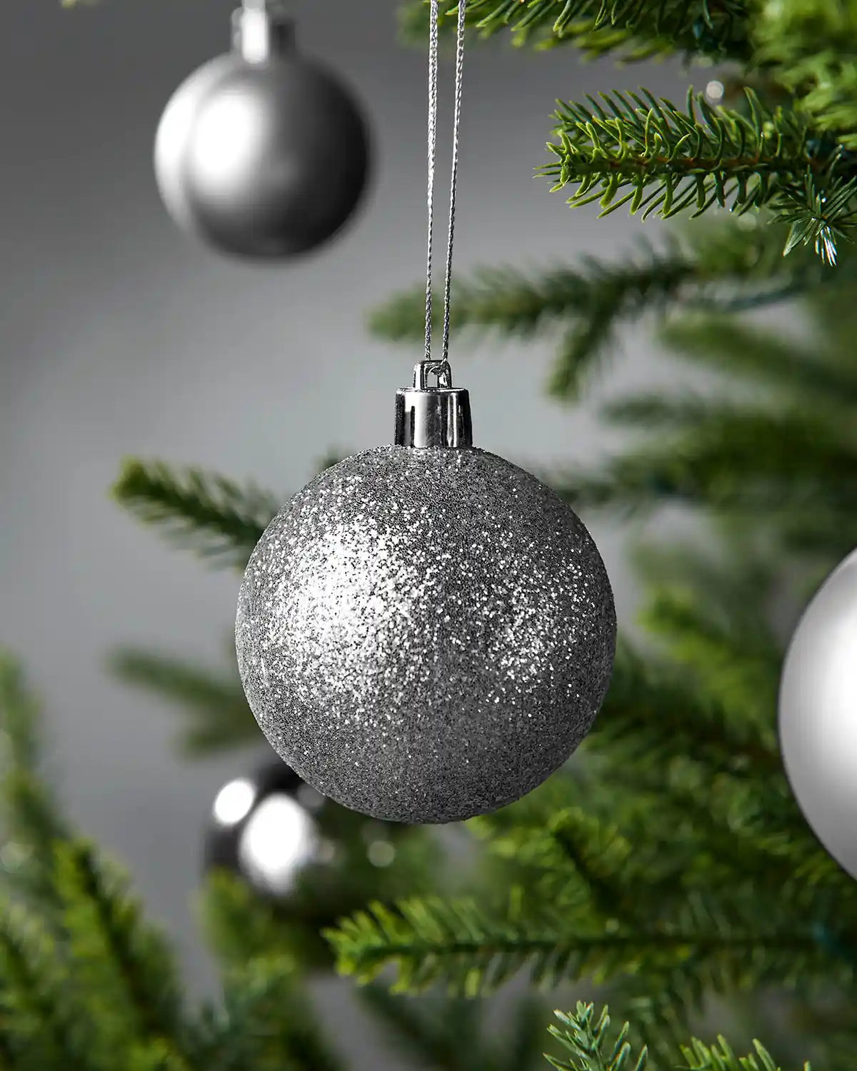 Close up of a small glitter silver bauble on a Christmas tree with blurry baubles in the background against a grey wall