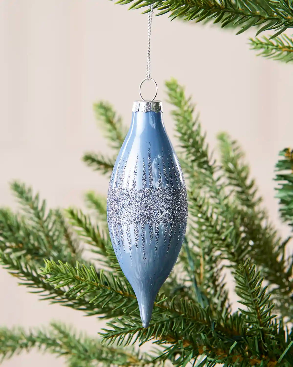 Close up of a teardrop shaped Ice blue glass bauble with glitter detailing, hanging from a Christmas tree branch. 