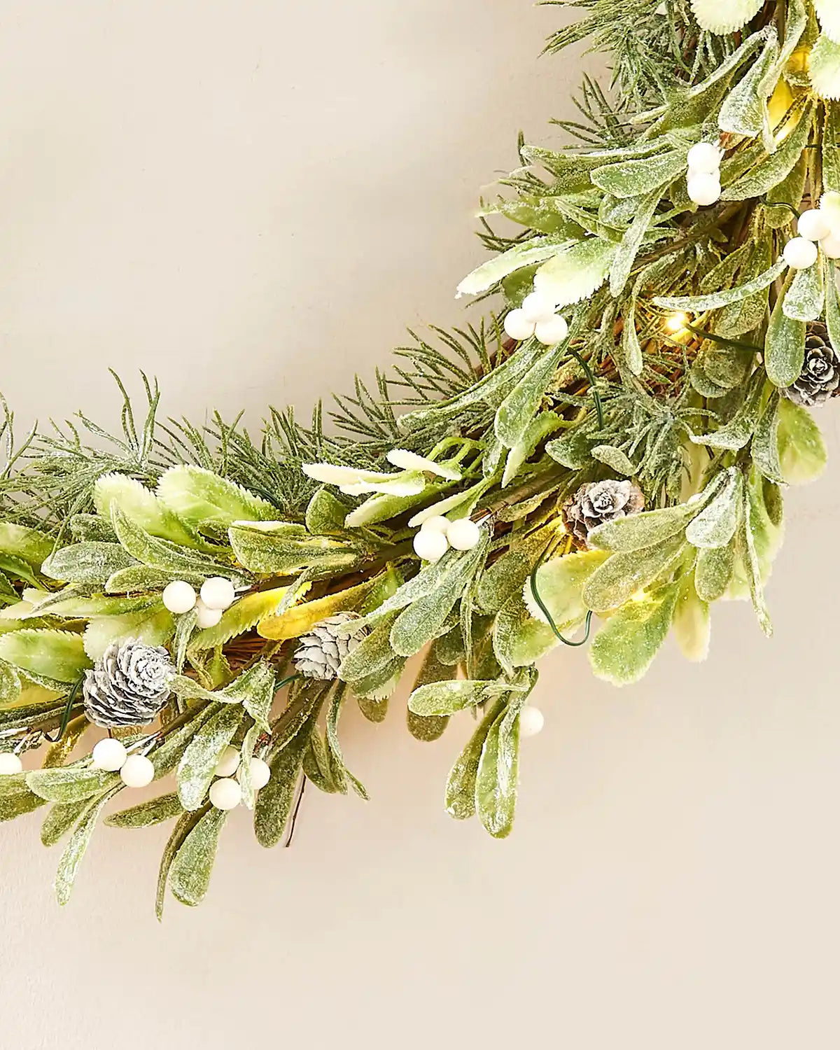 Close up of bottom of decorative wreath with greenery, pinecones, and small lights on a beige background