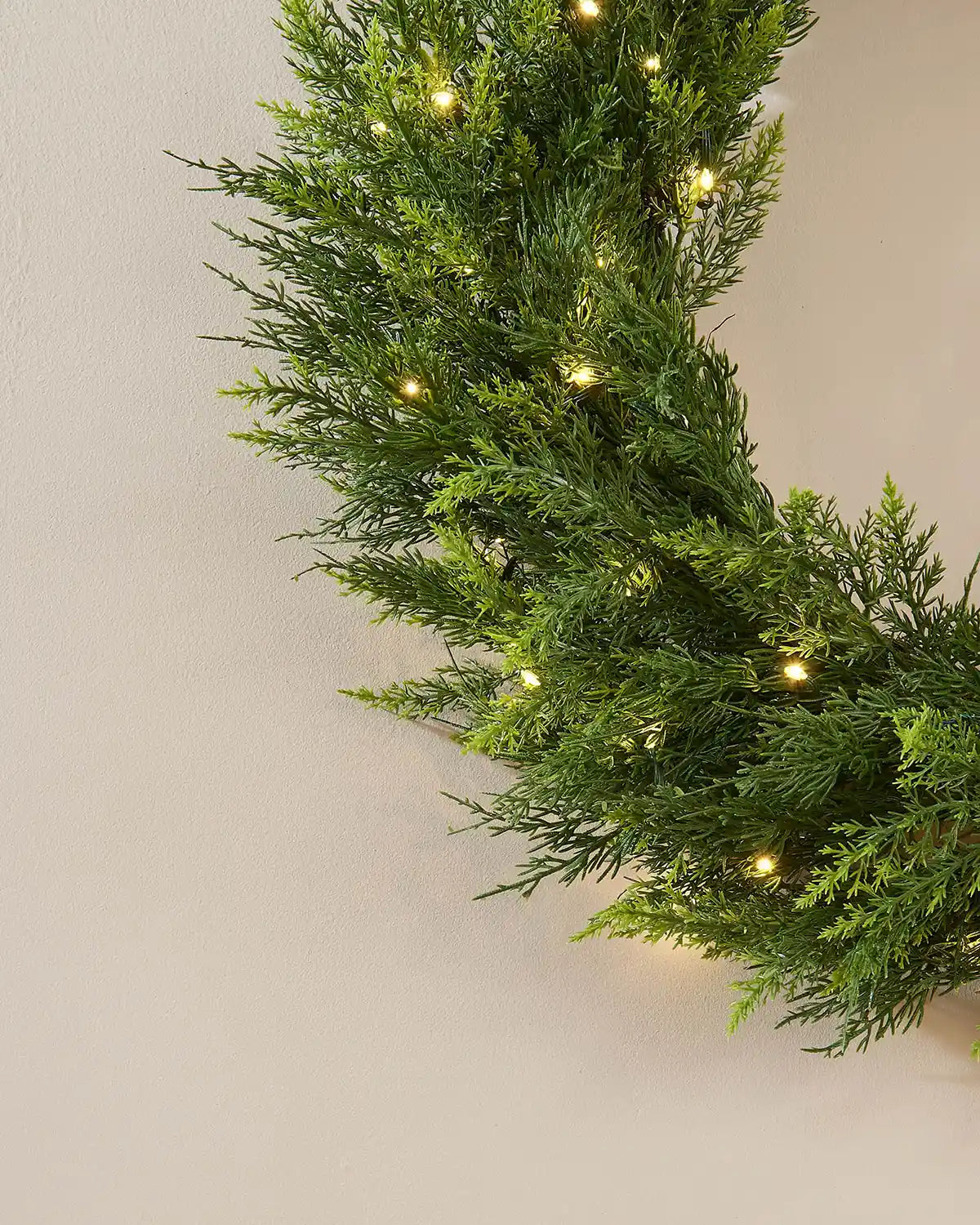 Close up of bottom of full looking green wreath with warm white lights on a cream wall