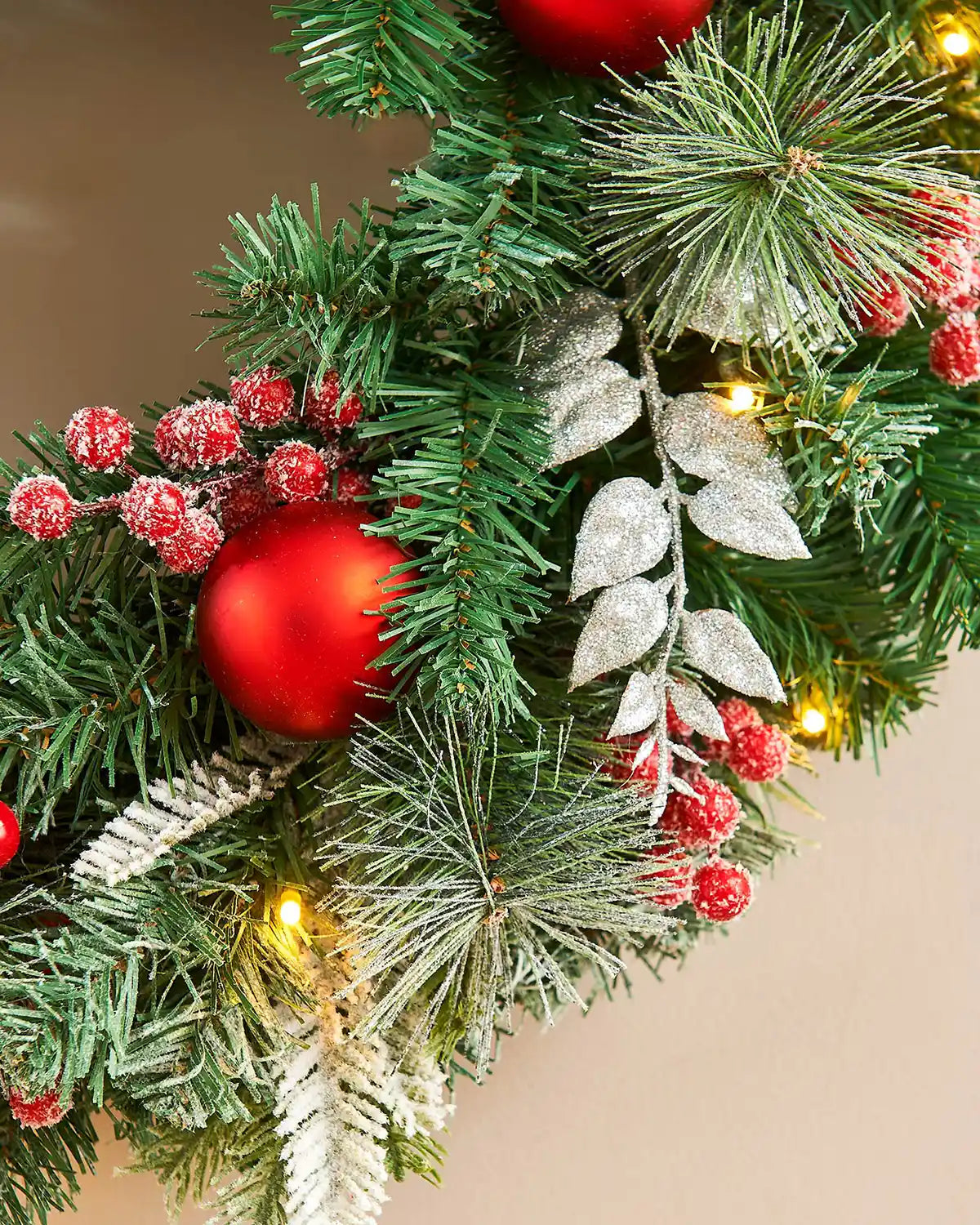 Close up of decorative Christmas wreath with red ornaments, berries, and lights on a neutral background