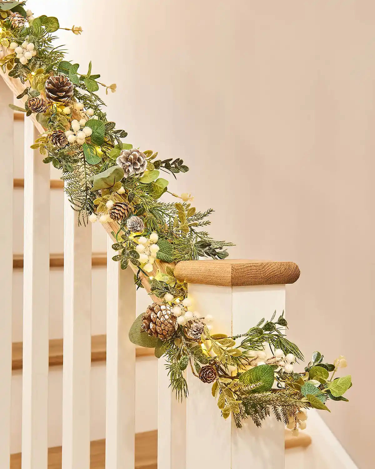 Close up of decorative green wreath and garland with White berries, pinecones and lights on a staircase banister with a neutral background