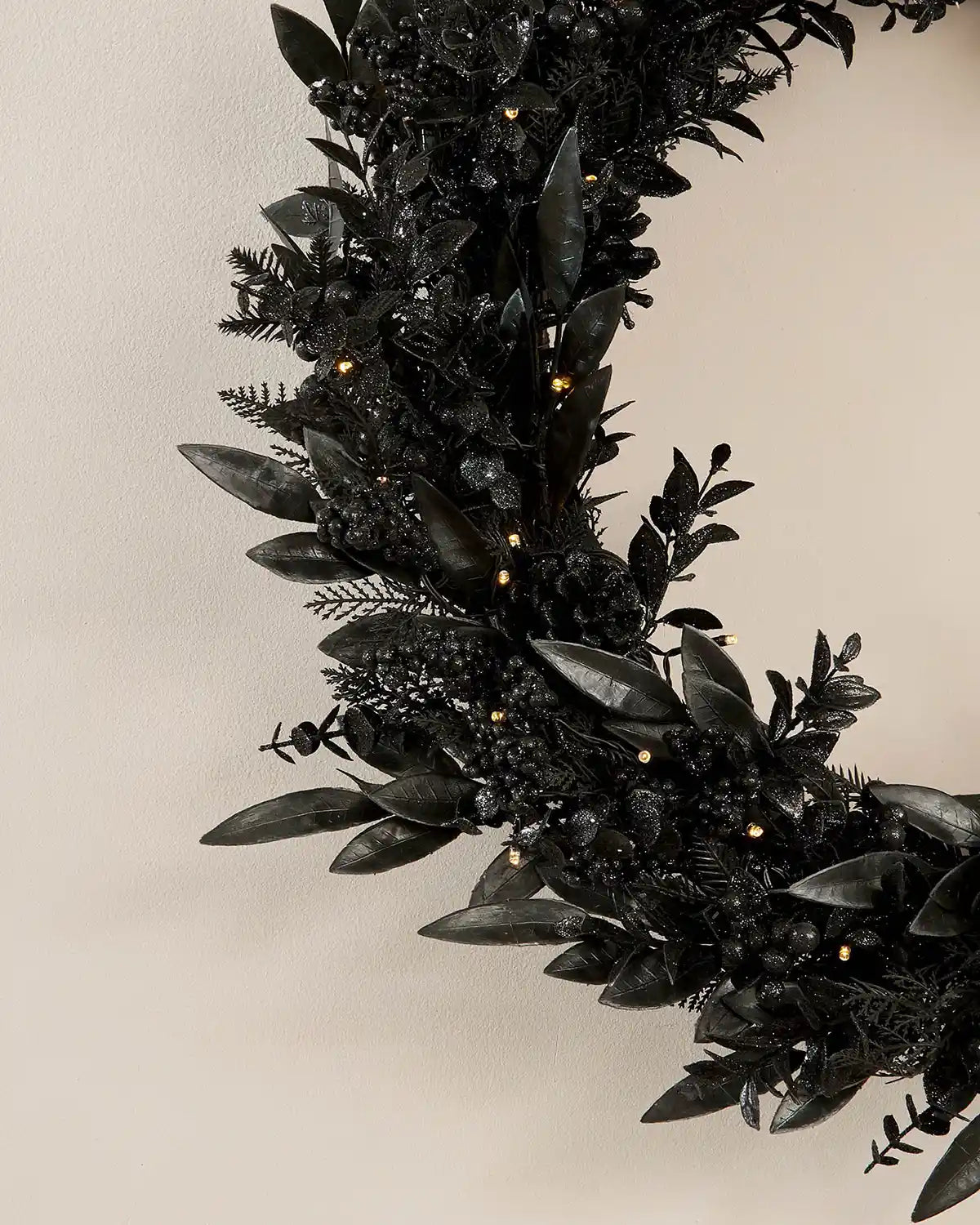 Close up of side of Black decorated wreath with black pine cones, berries and warm white lights against a cream wall