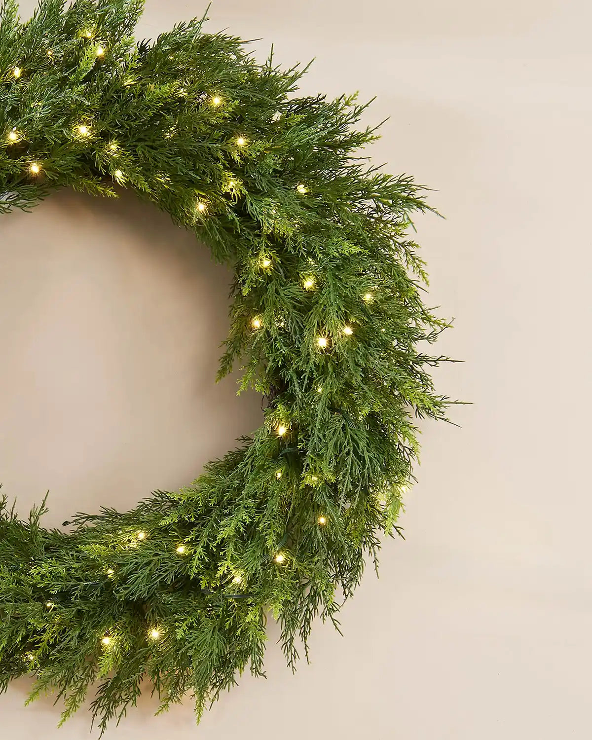 Close up of side of full looking green wreath with warm white lights on a cream wall