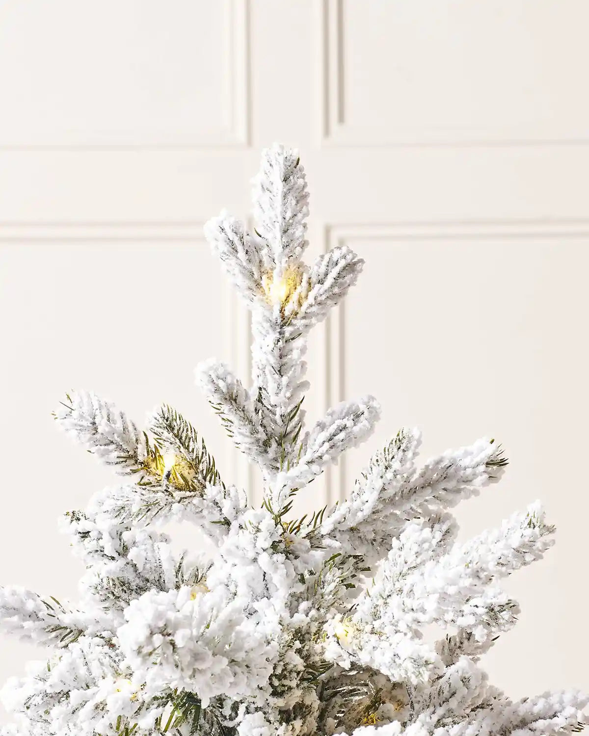 Close up of the top of a Pre-Lit Snow Flocked Potted Christmas Tree showing the Snow, single lit bulbs against a cream panel wall