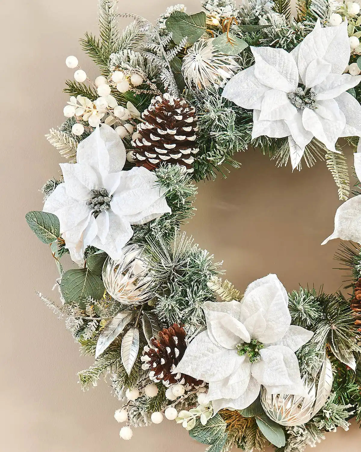 Close up of wreath with white flowers, pinecones, and greenery on a beige background