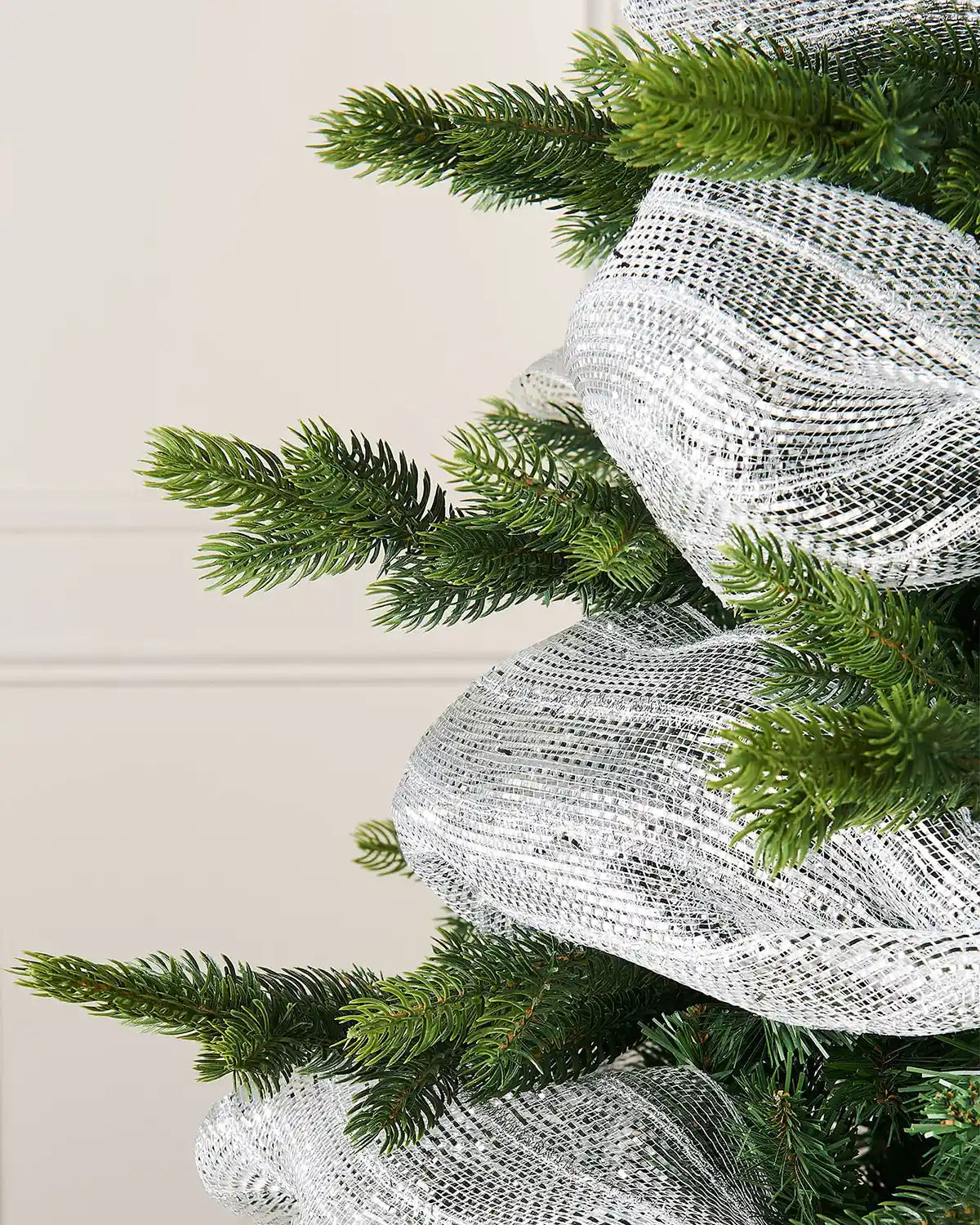 Close up transparent and silver foil mesh fabric ribbon wrapped around Christmas tree against a cream panel wall