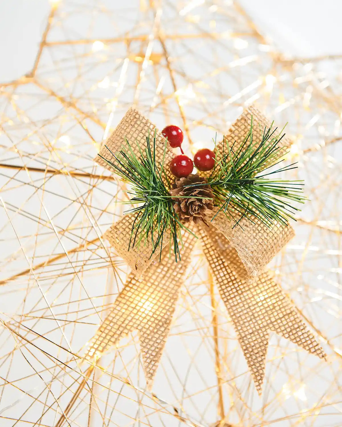 Close up view of the Golden bow with Mistletoe, berries and a pine cone part of  Pre-Lit 2D Metal Star, Gold, 60 cm on a White table with Pinecone lantern in front of a staircase with lights and a garland 