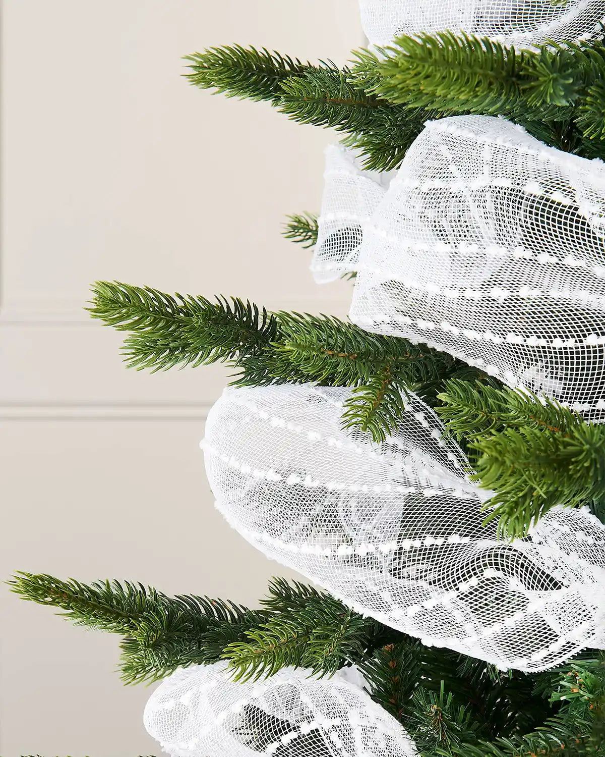 Close up white decorative mesh ribbon with lace pattern wrapped around Christmas tree against cream wall