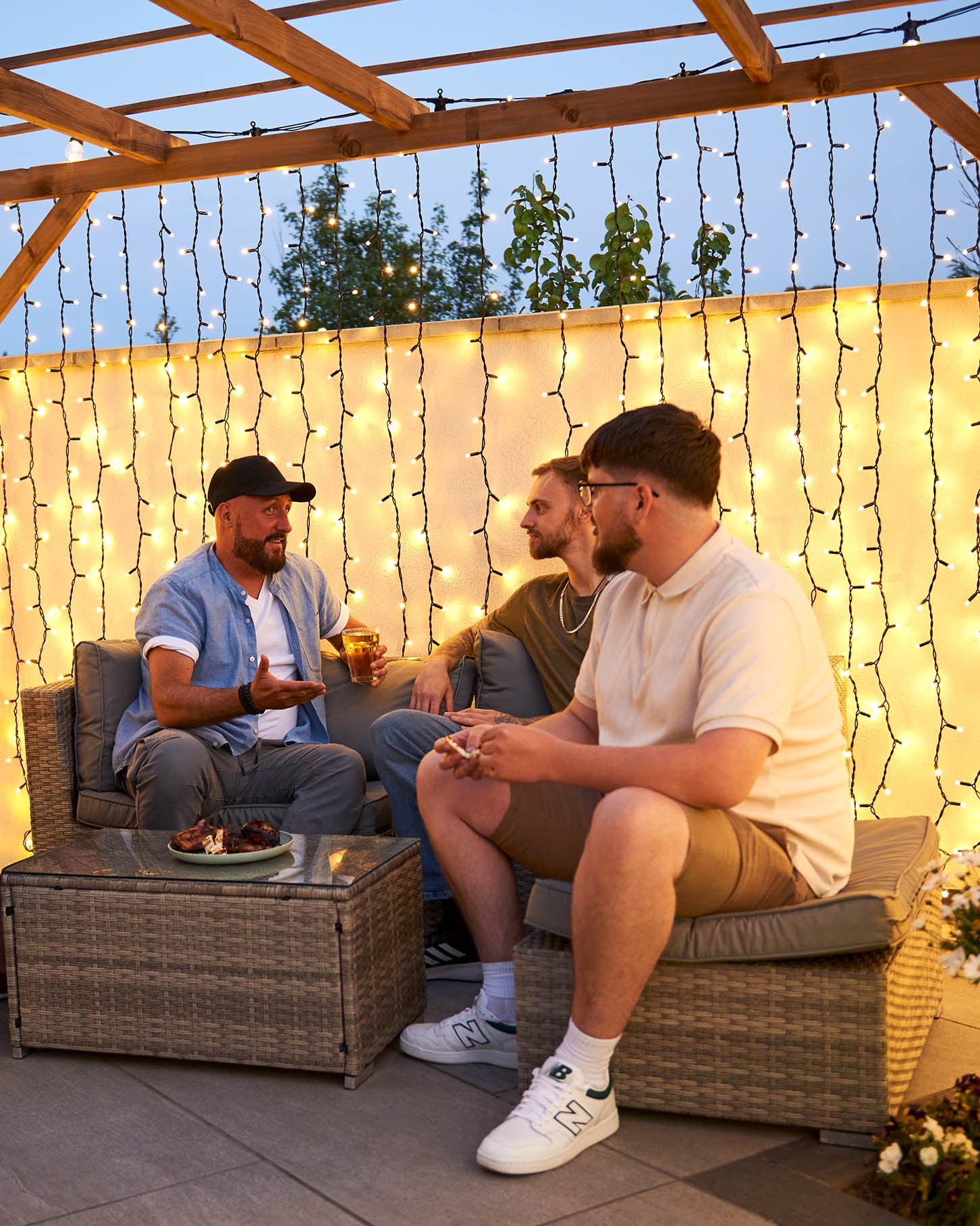 Three men sitting on outdoor furniture under curtain lights on a patio.