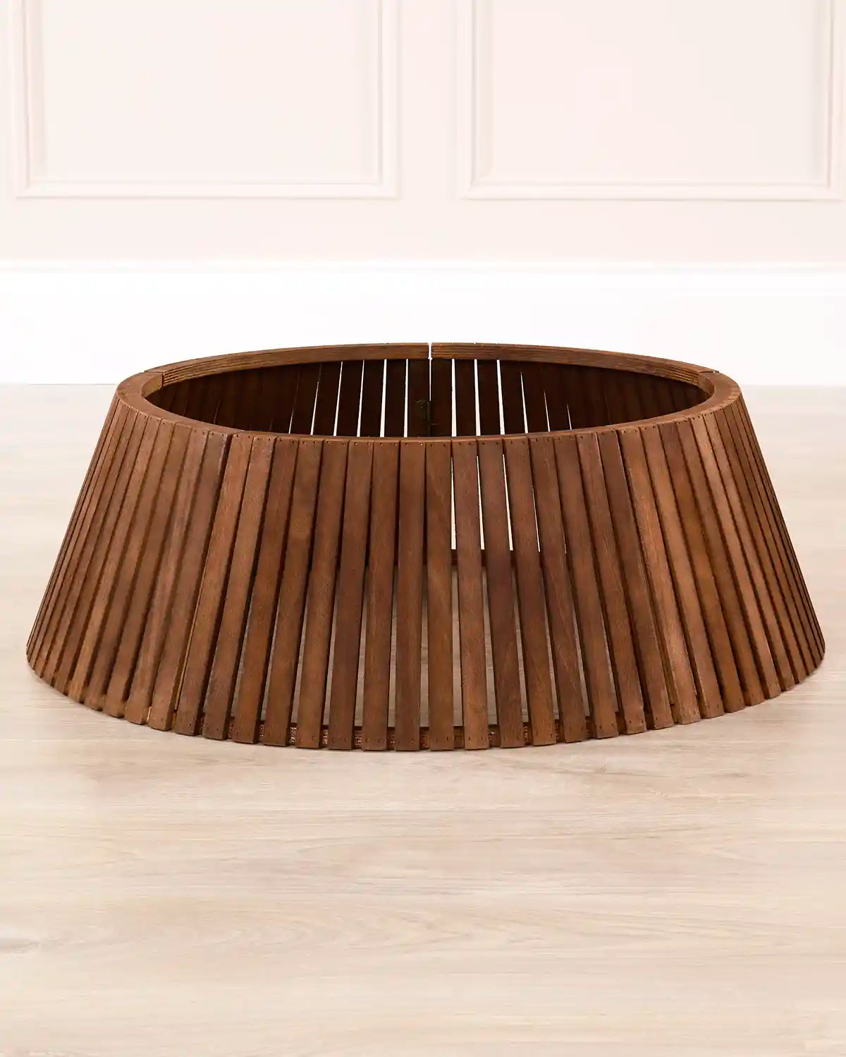 Dark brown Christmas tree collar made of vertically slatted wood in a medium brown finish, set on a light wood floor against a white panelled wall.