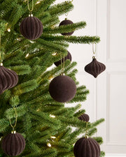 Dark brown velvet Christmas tree ornaments hanging on a green pine tree with warm white string lights, set against a white paneled wall.